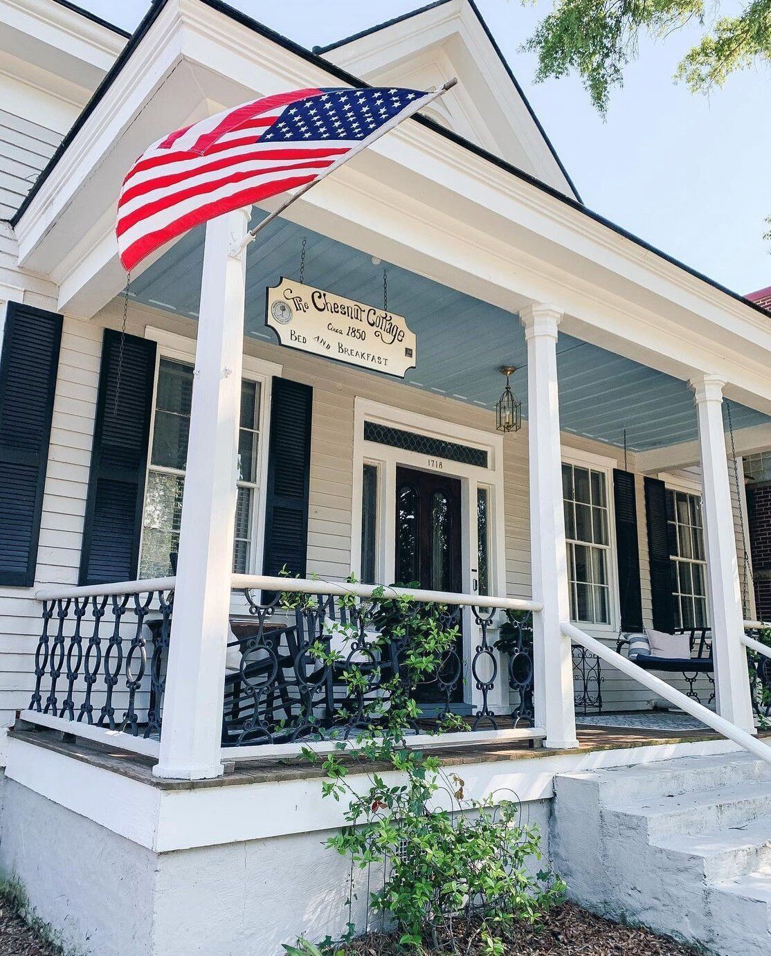 White house with porch, American flag, black shutters, blue ceiling, and decorative railing.