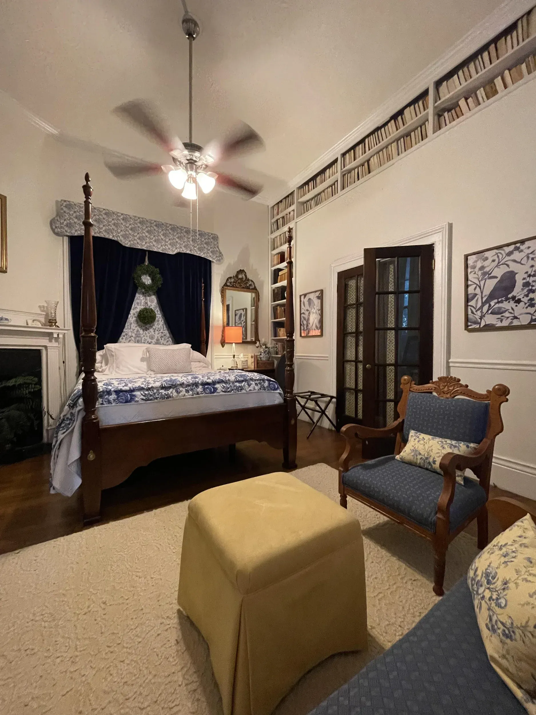 Cozy bedroom: Four-poster bed, built-in bookshelves, blue and white decor, armchair, ottoman, and French doors.