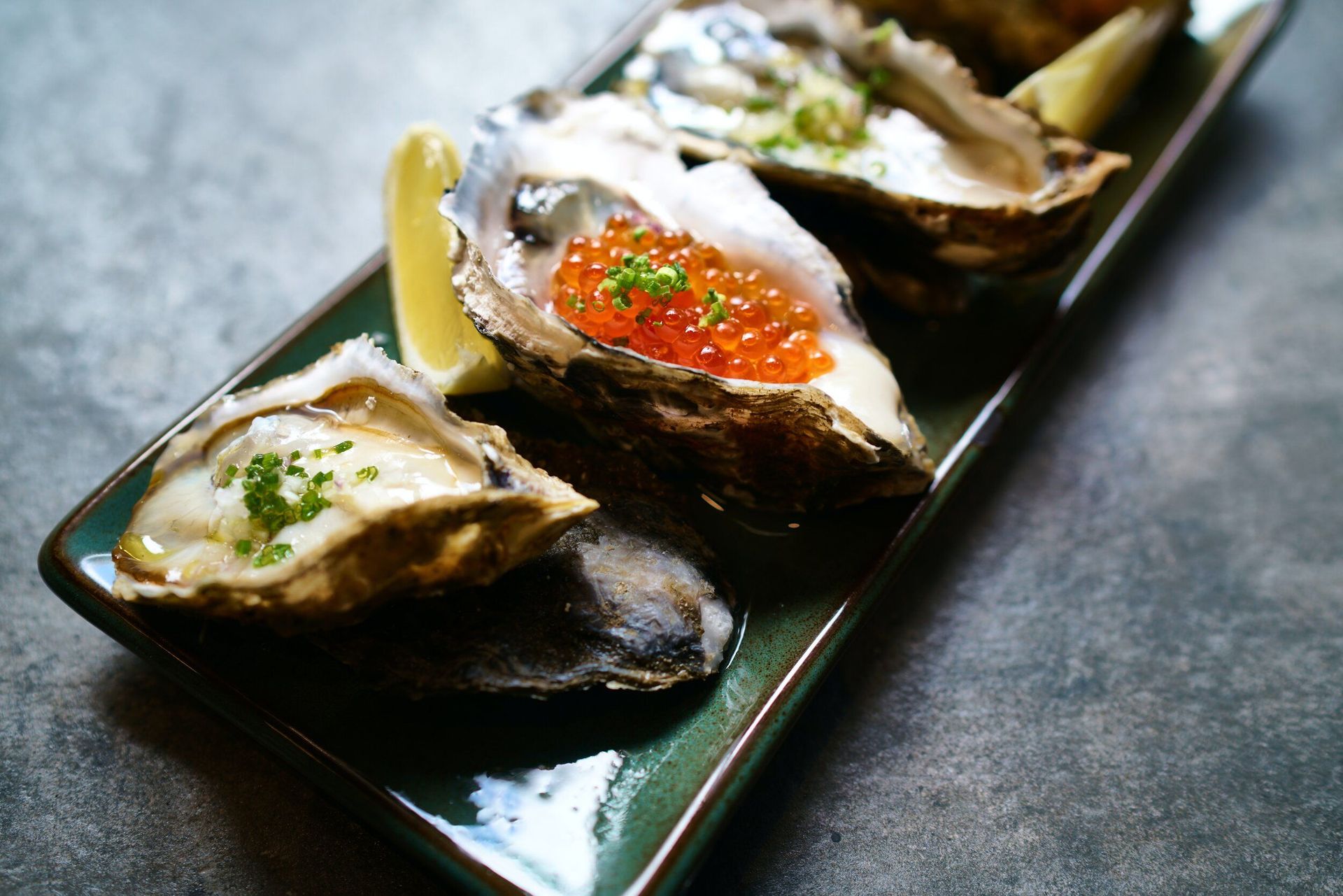 Oysters on a green plate, topped with roe, herbs, and lemon wedges, set against a gray background.
