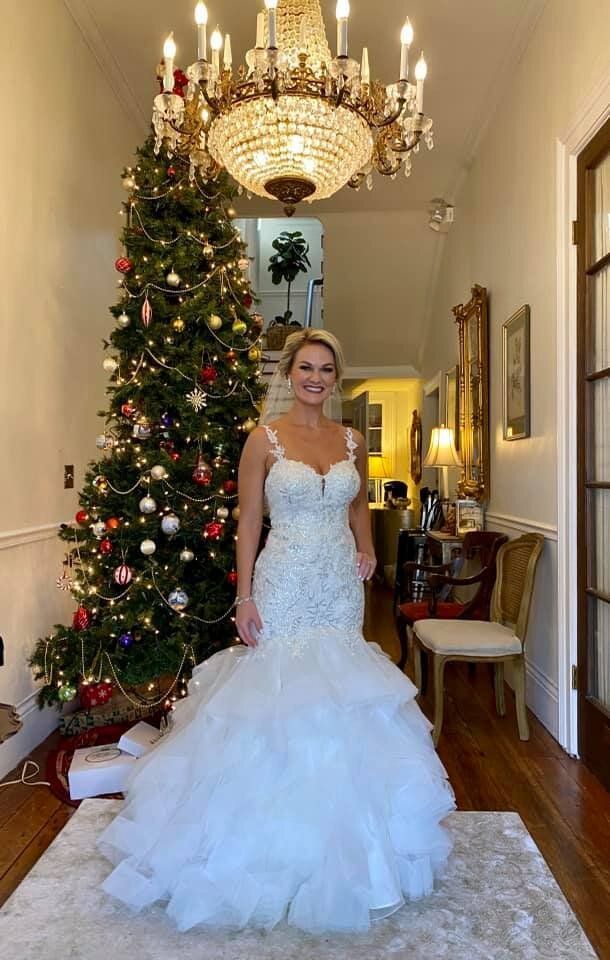Woman in a white wedding dress stands by a Christmas tree under a chandelier in a hallway.