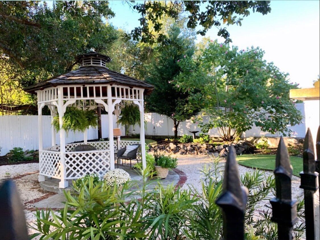 White gazebo in a garden with seating, greenery, and a brick pathway. A black fence is in the foreground.