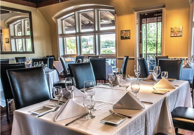 Restaurant dining room with tables set for service; large windows, mirrors, dark chairs, and white tablecloths.