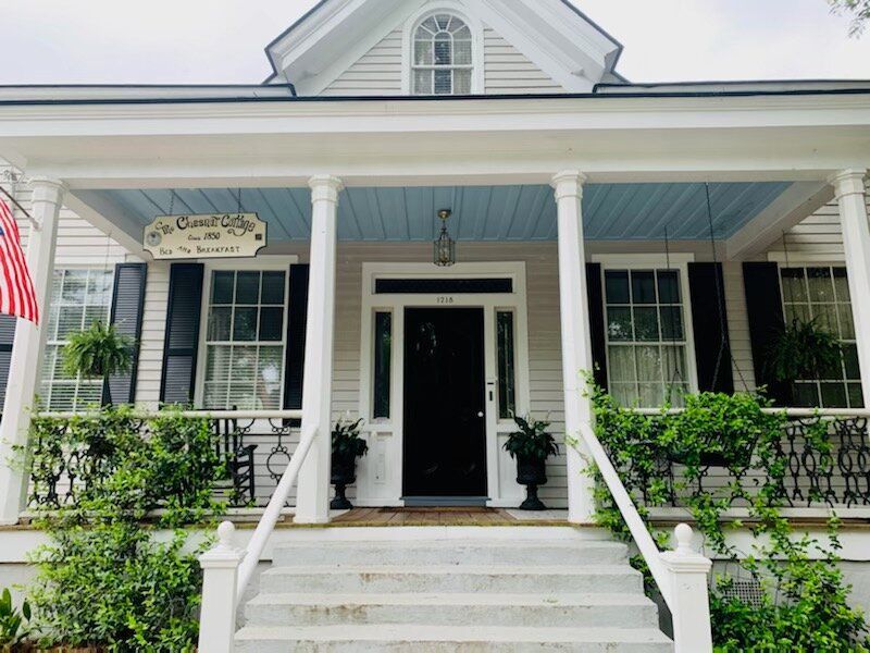 Front porch of a two-story house with white trim and blue ceiling. Black door, steps, and lush greenery.