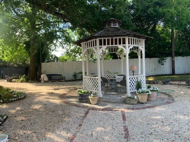 White gazebo in a gravel yard with potted plants and a brick path, shaded by a large tree.