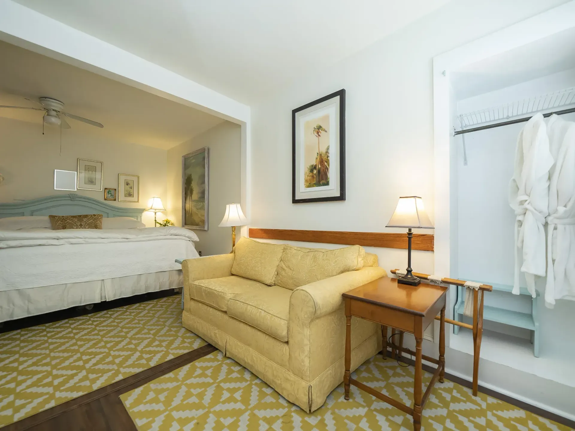 Cozy hotel room with a yellow couch, small table, and an open doorway to a bed. A closet with robes is on the right.
