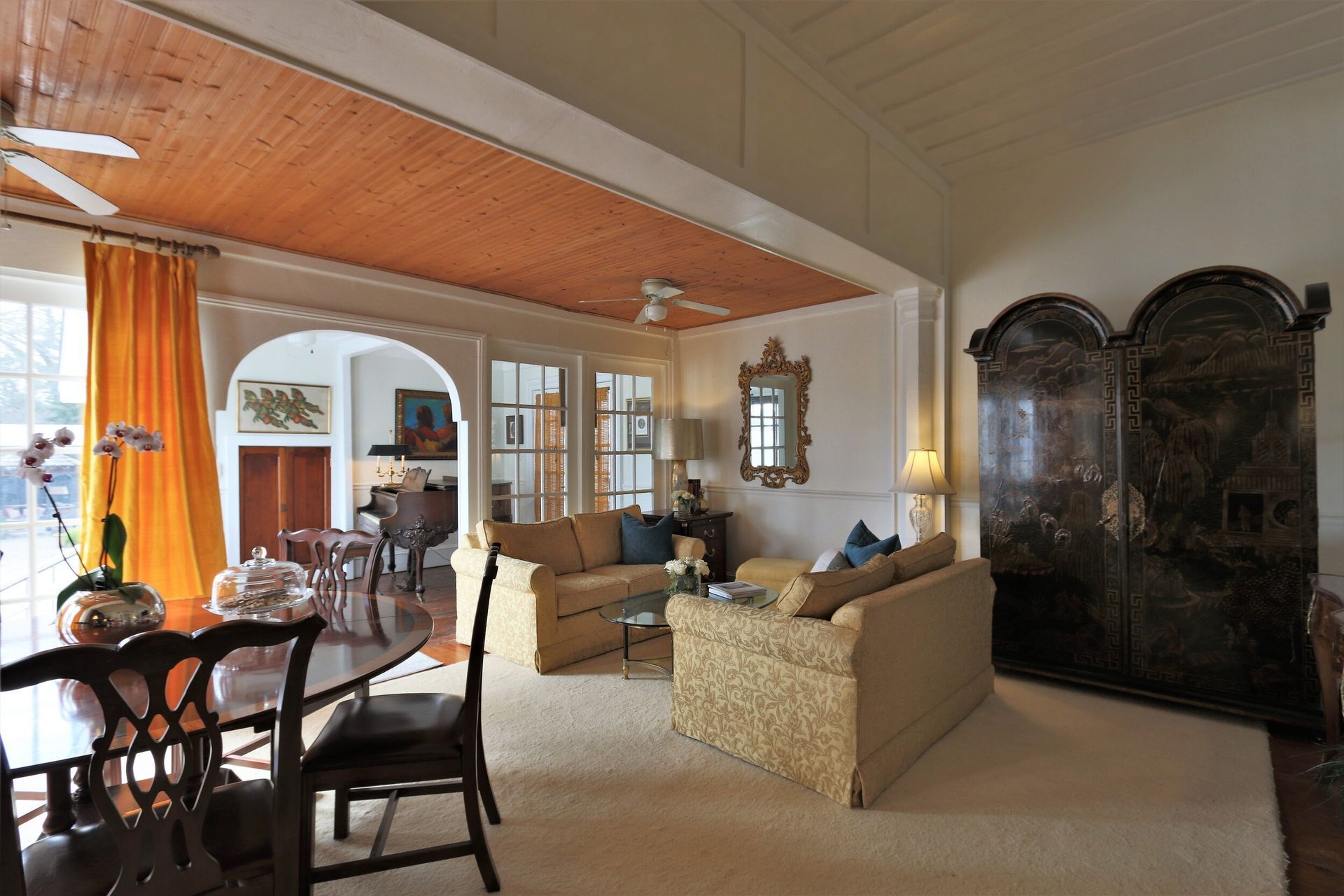 Spacious living room with dining area, orange curtains, light-colored sofas, and ornate wooden storage cabinet.