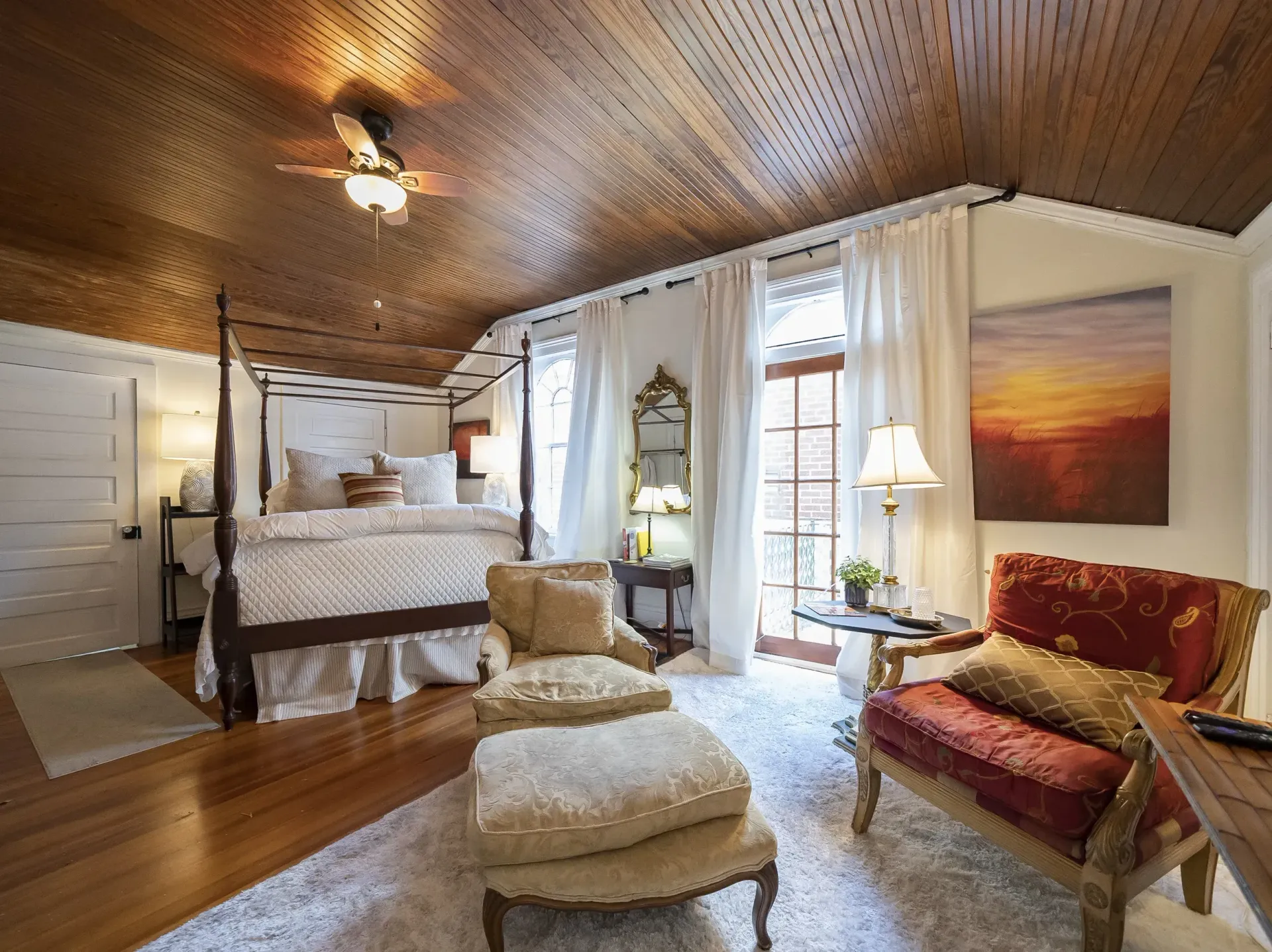 Bedroom with four-poster bed, ornate chair, ottoman, white curtains, and a wood-paneled ceiling.