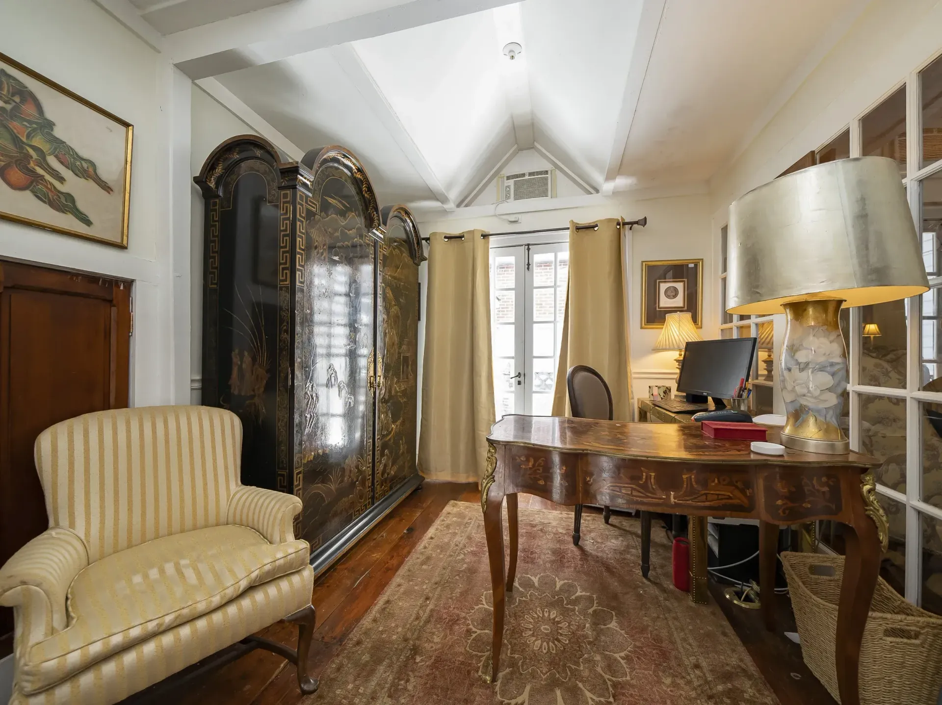 Cozy office: antique desk, patterned rug, gold-toned lamps, armchair, ornate screen, window with curtains, and artwork.