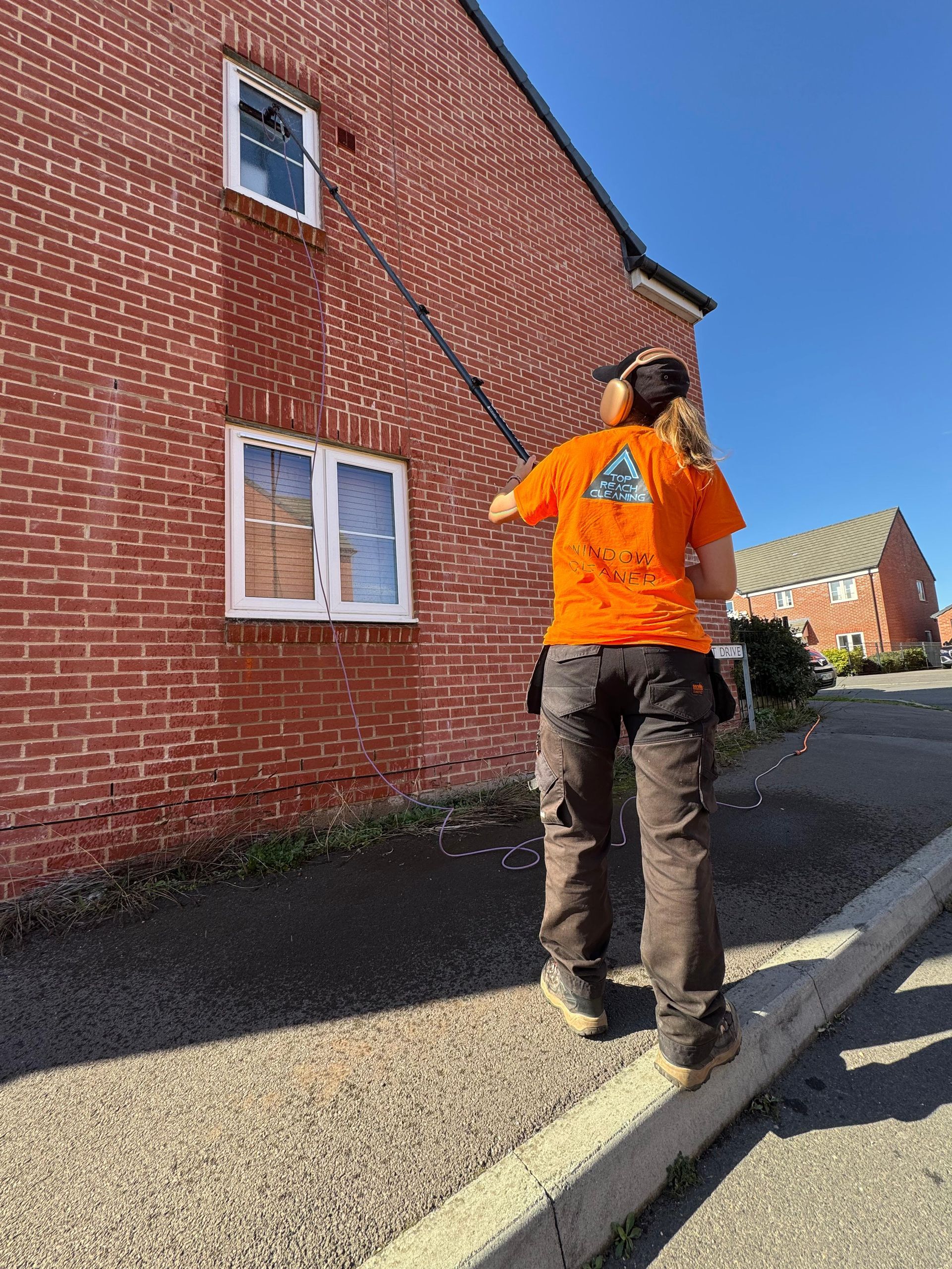 Professional window cleaner on a ladder washing second-floor windows of a suburban house under clear