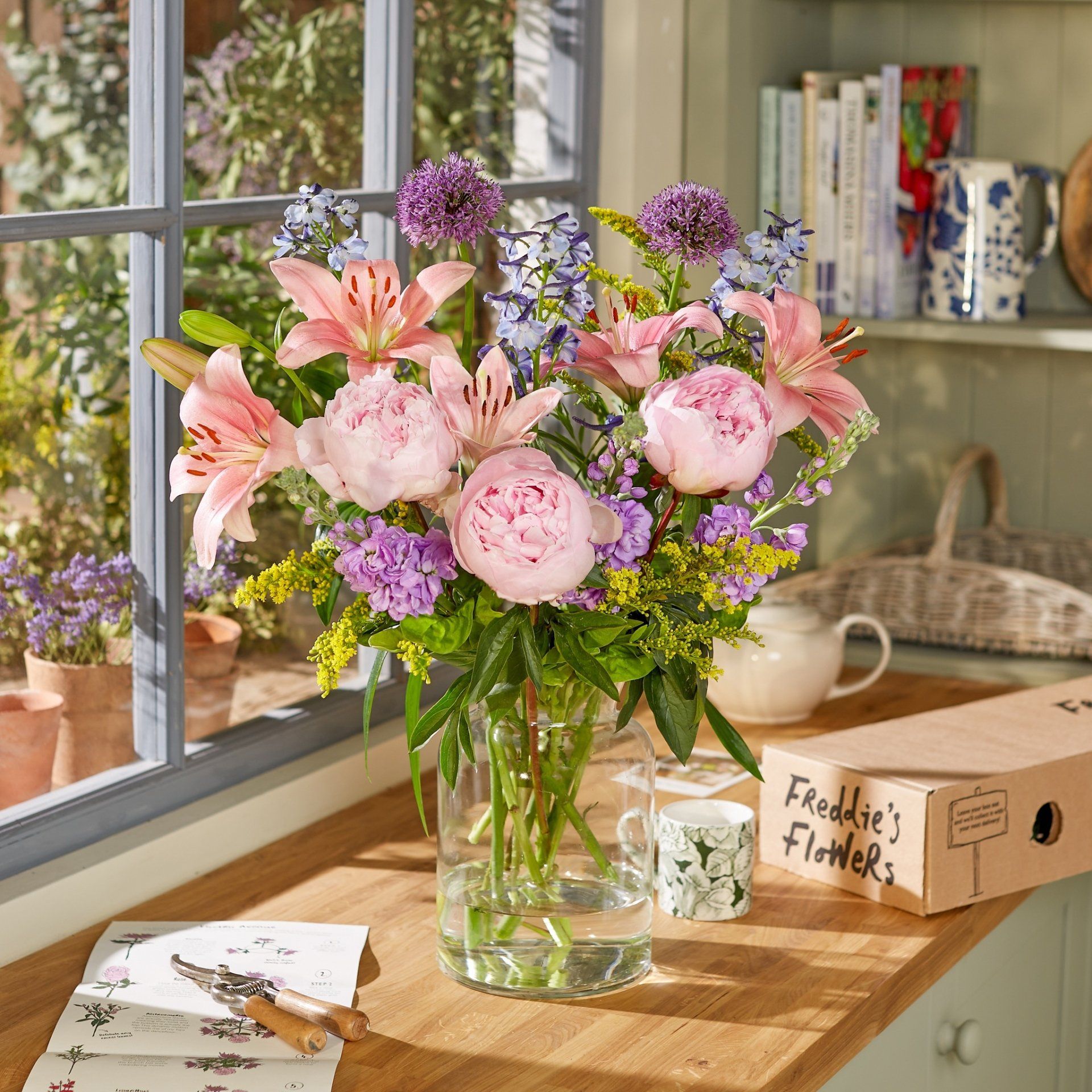 A vase filled with pink and purple flowers sits on a table next to a box that says feedde 's flowers