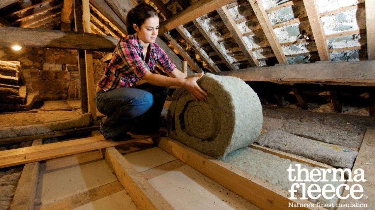 A woman is kneeling down in an attic holding a roll of insulation.