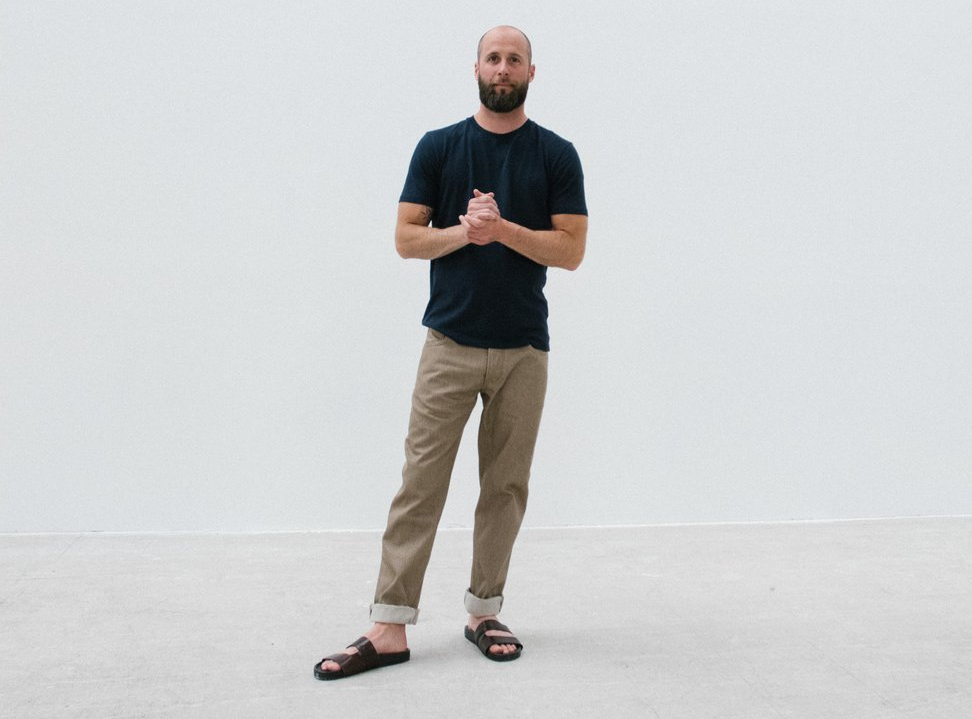 A bald man with a beard is standing in front of a white wall with his hands folded.