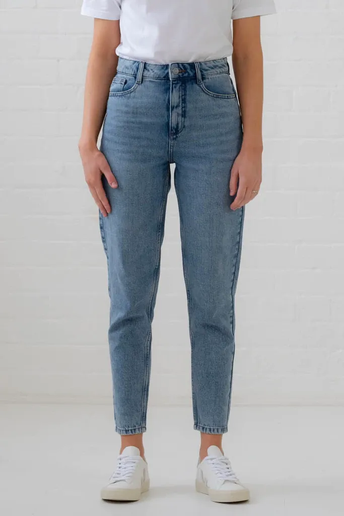 A woman wearing a white shirt and blue jeans is standing in front of a white wall.