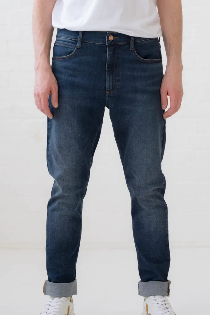 A man wearing a white shirt and blue jeans is standing in front of a white wall.