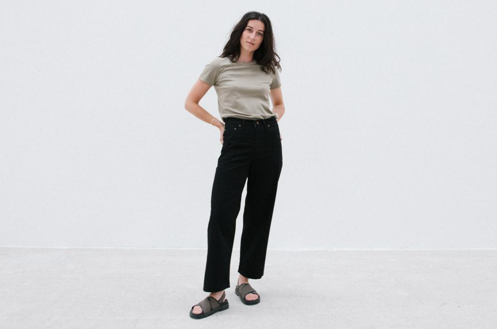 A woman wearing a t-shirt and black pants is standing in front of a white wall.