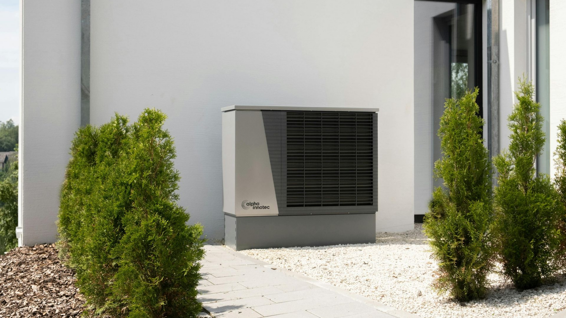 Heat pump unit outside a building, surrounded by greenery. Gray and black.