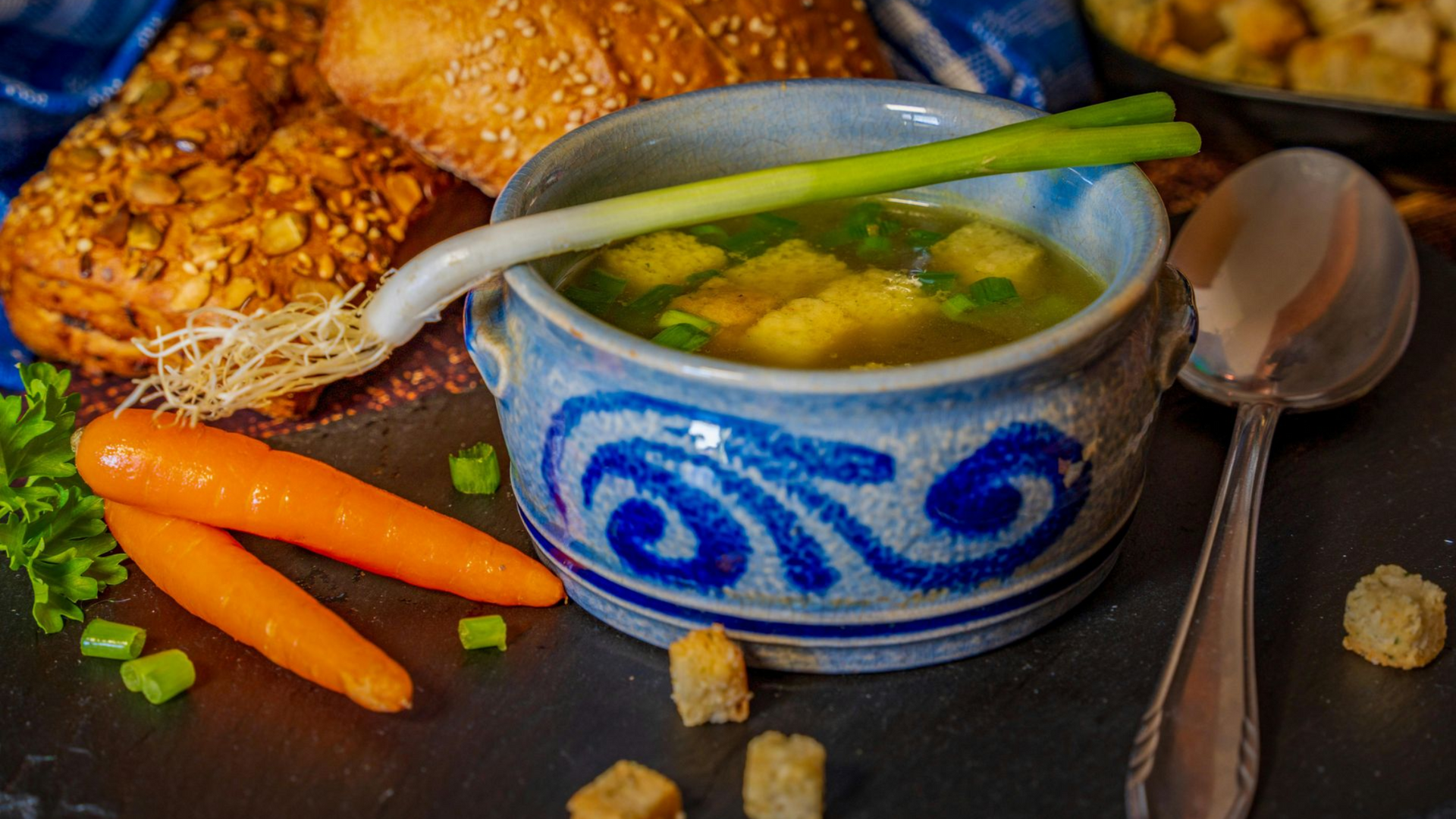 Soup in a blue bowl with bread, carrots, and green onions on a slate.