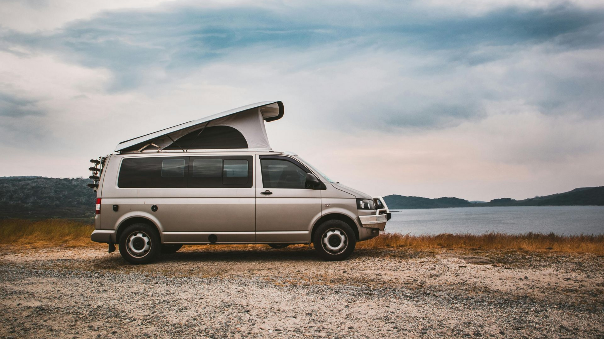Tan campervan parked on gravel near a lake and trees under a cloudy sky.