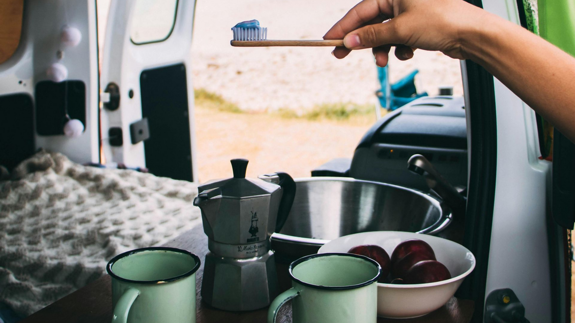 Person holding a toothbrush with blue paste in a campervan. Coffee pot, mugs, bowl of fruit visible.