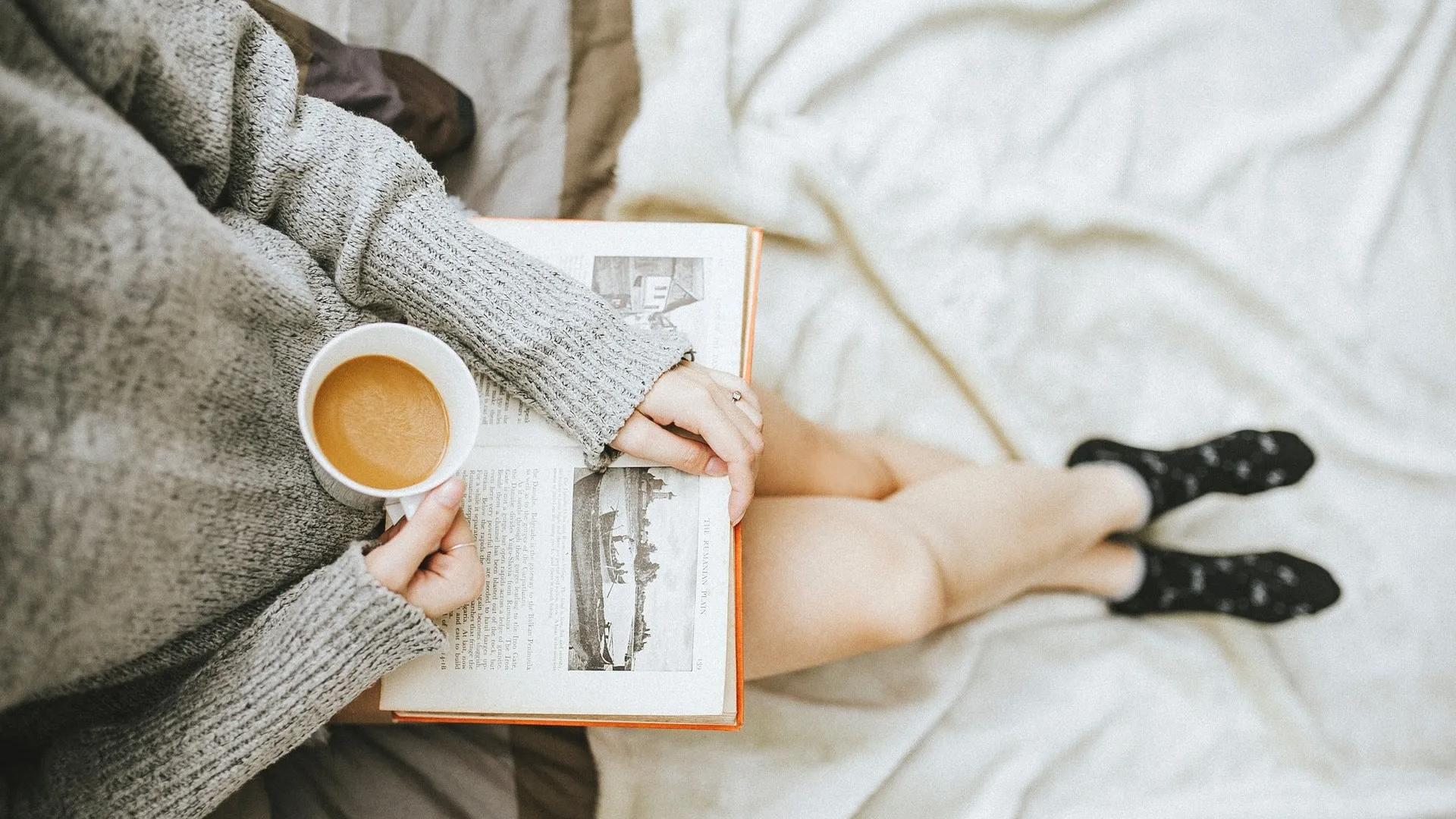 A woman is sitting on a bed reading a book and drinking coffee.