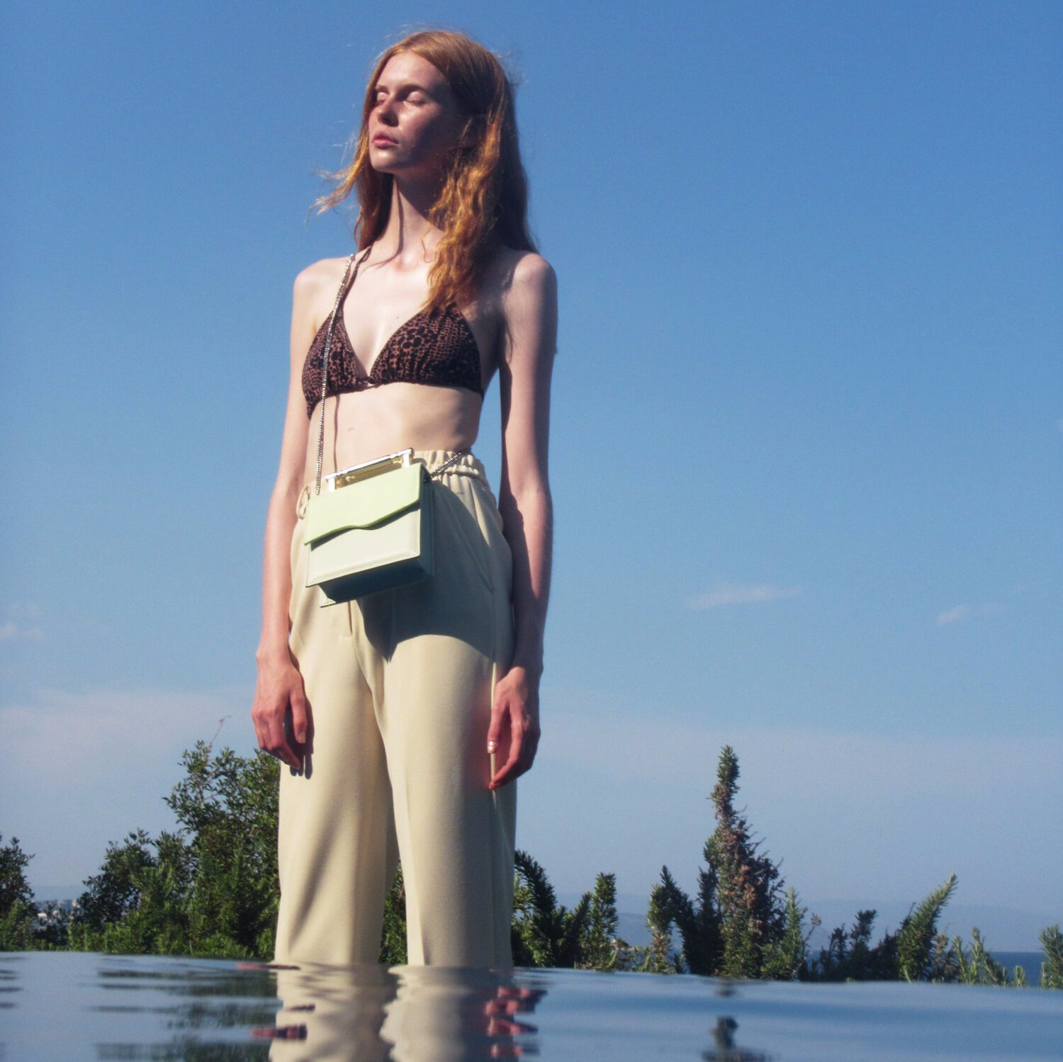 A woman in a bikini top and pants is standing in the water wearing a luxury bag