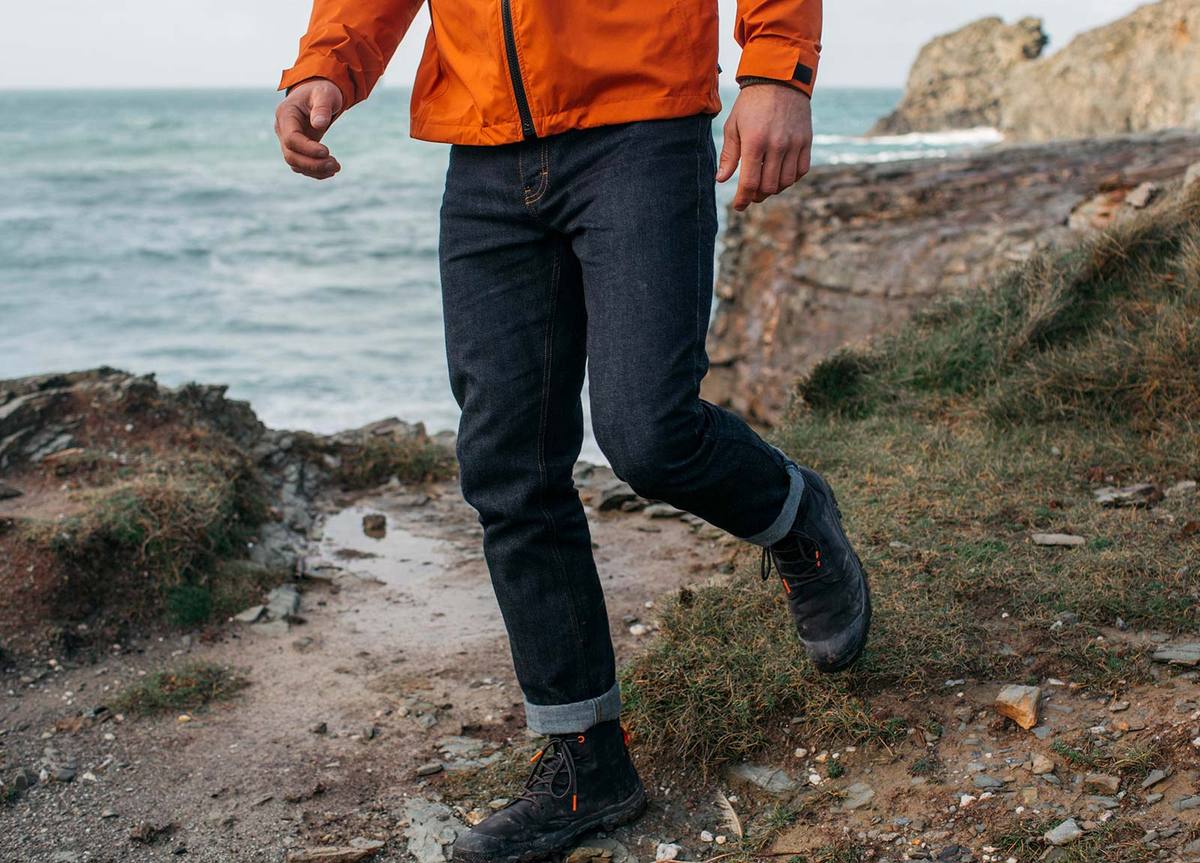 A man in an orange jacket and jeans is running on a rocky beach near the ocean.