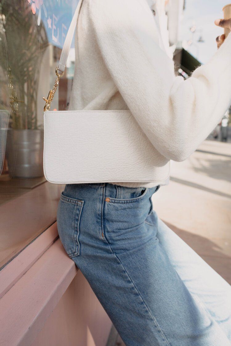 A woman wearing a white sweater and blue jeans is holding a white bag.