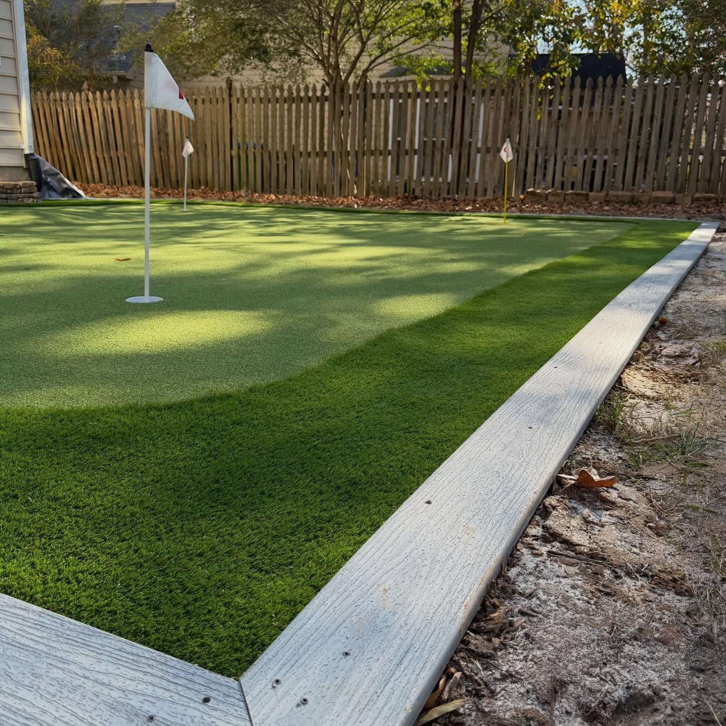 Artificial turf putting green with a flagstick and white border in a fenced backyard