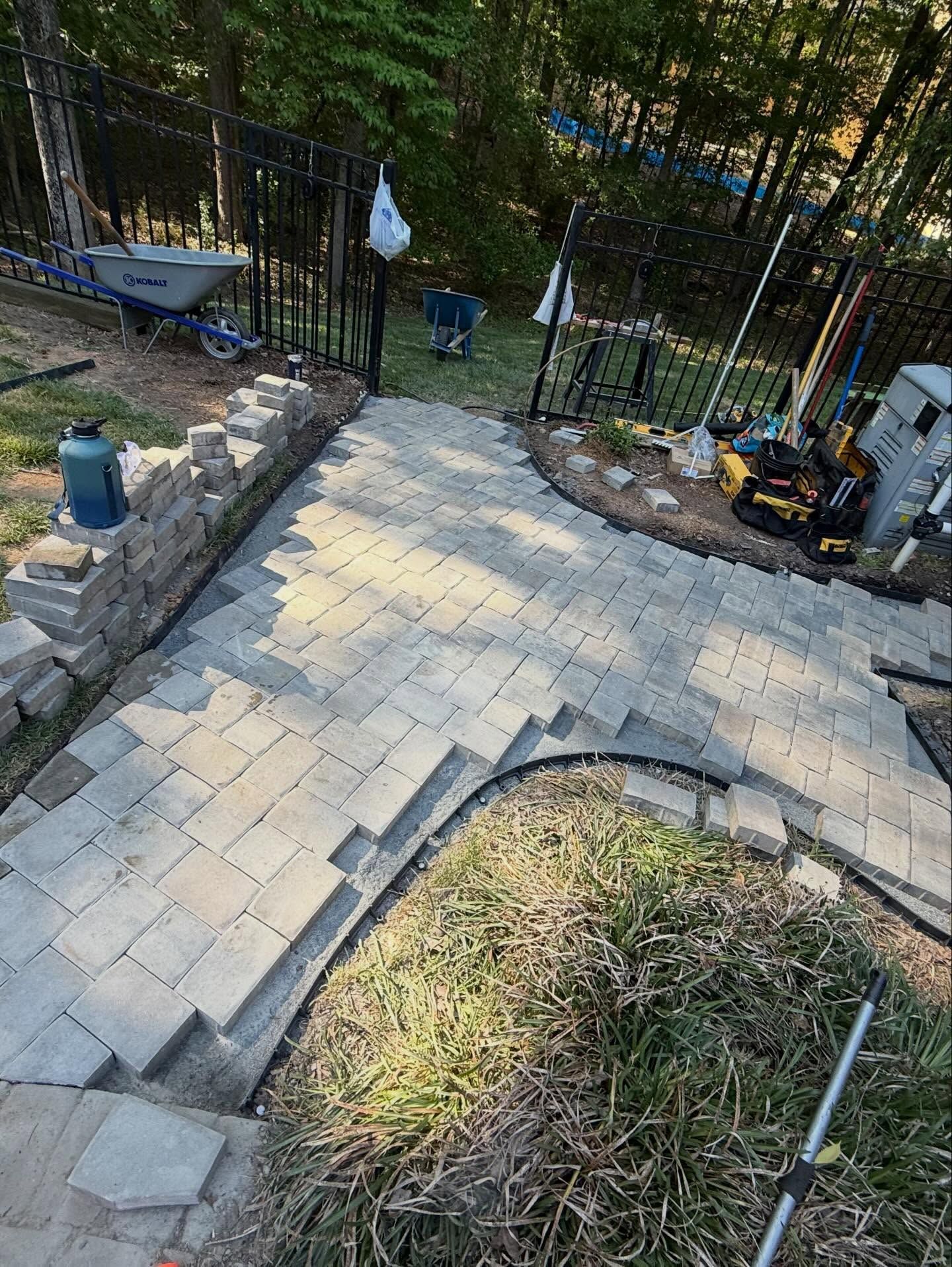 Stone paver walkway leading down a sloped backyard toward a black gate and wooded area