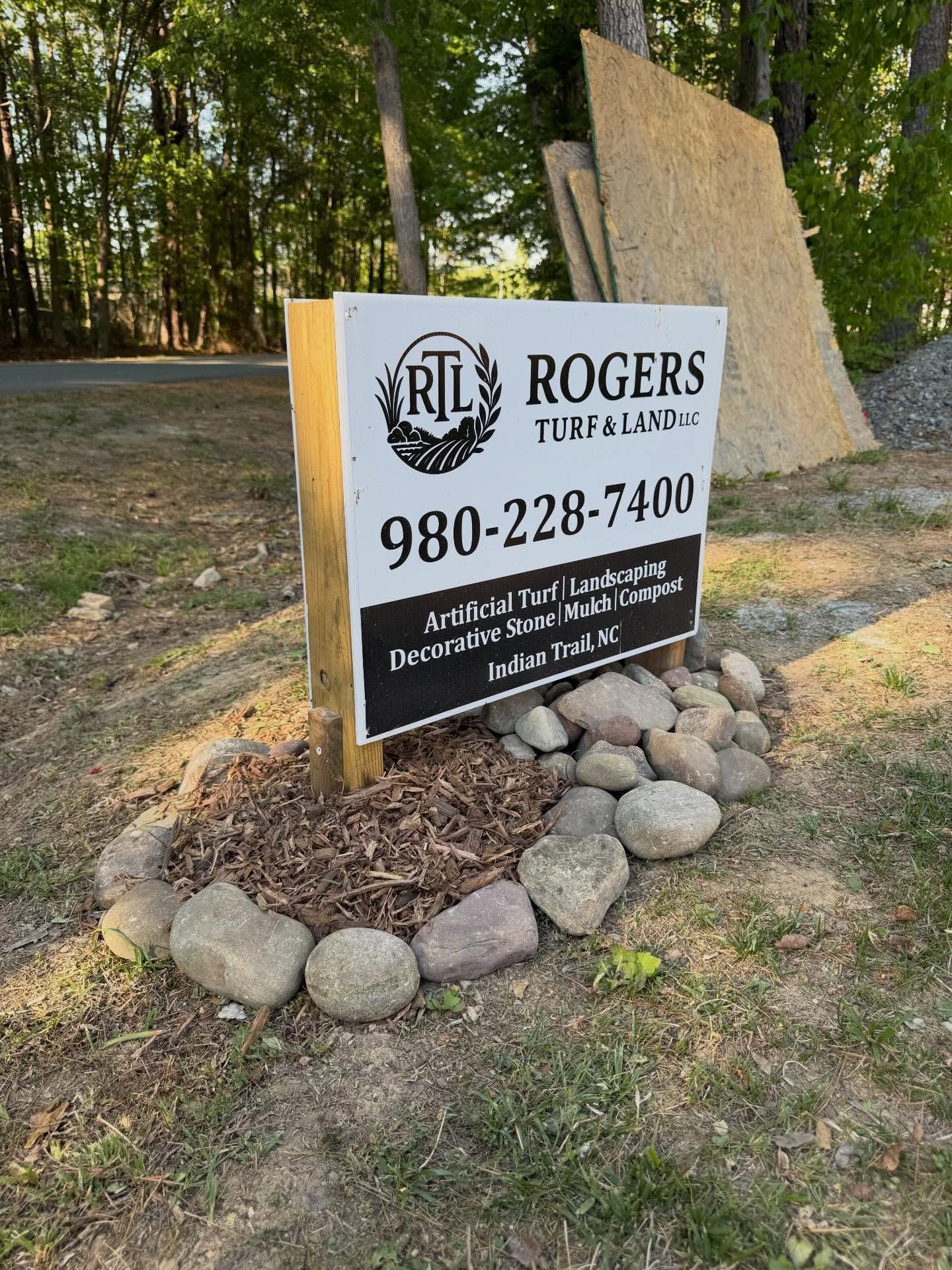 Rogers Real Estate sign with phone number, surrounded by rocks in a wooded outdoor setting