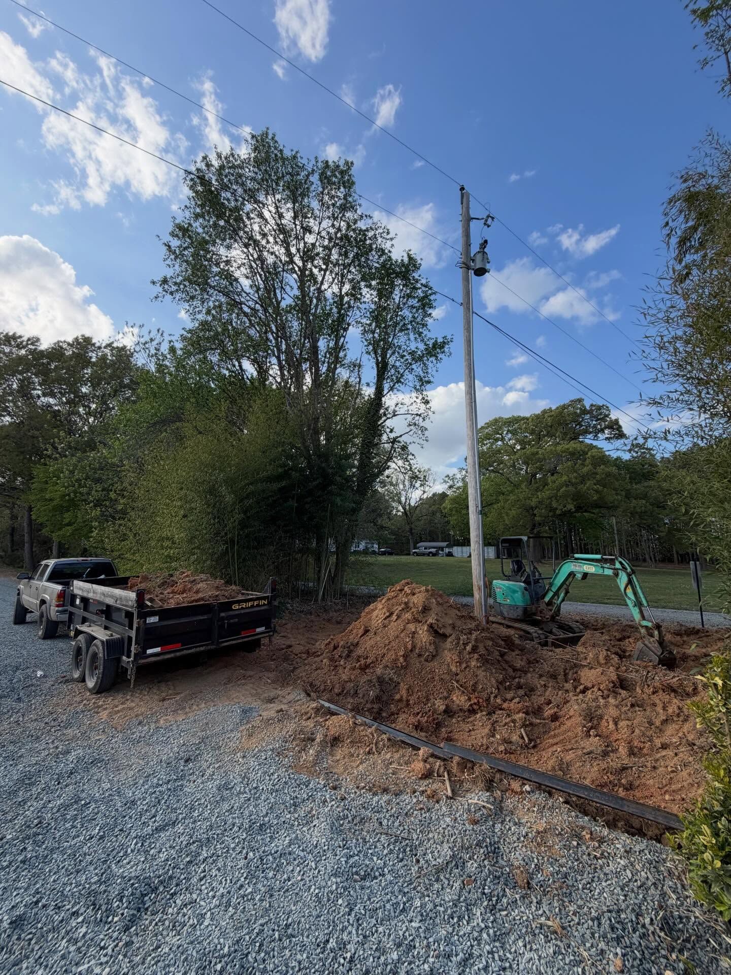 Construction site with excavator, dump trailer, and gravel beside a tall tree under a blue sky