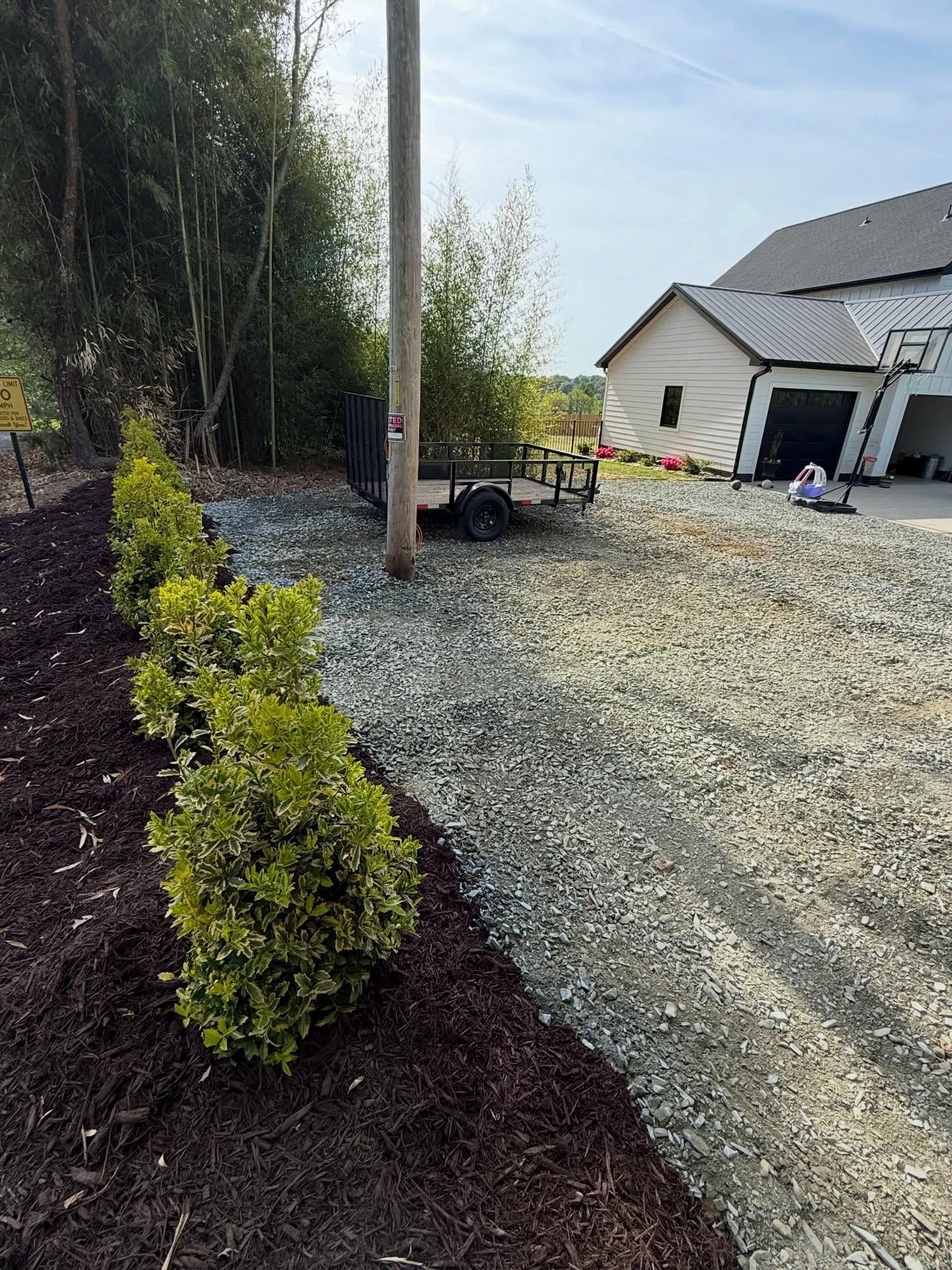 Gravel driveway beside a white house with a detached garage and a row of small shrubs along a mulch bed.