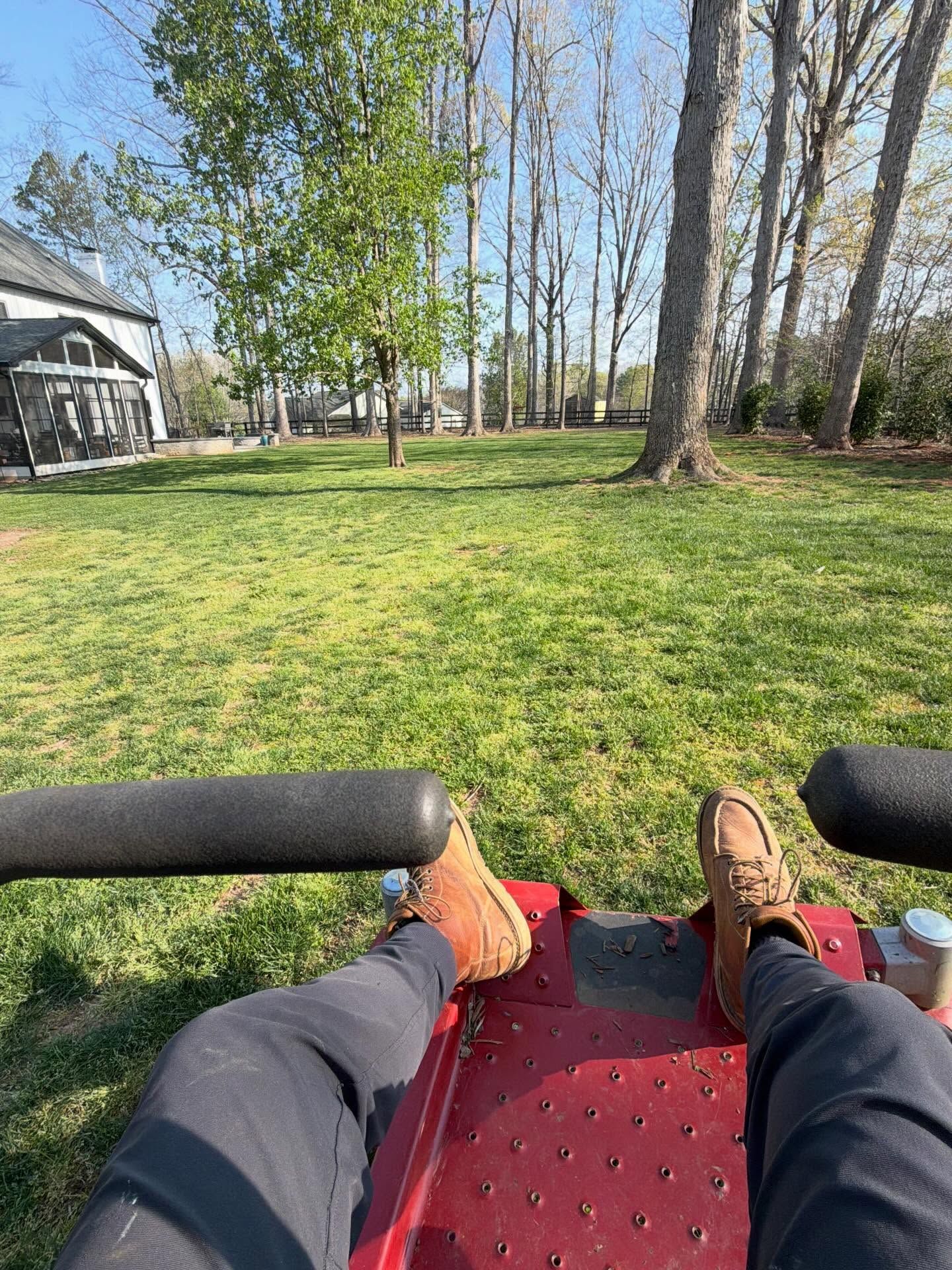 First-person view of feet on a red riding mower overlooking a grassy yard and trees