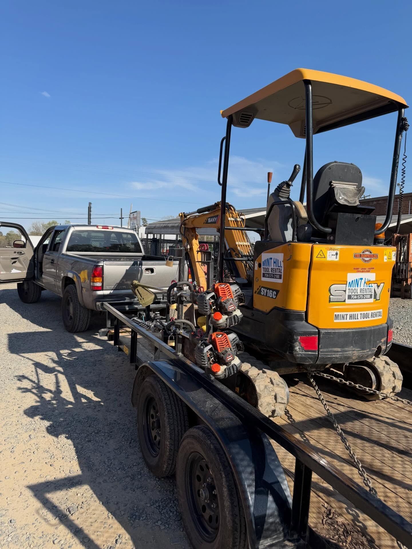 Yellow mini excavator on a trailer behind a pickup truck on a dirt road