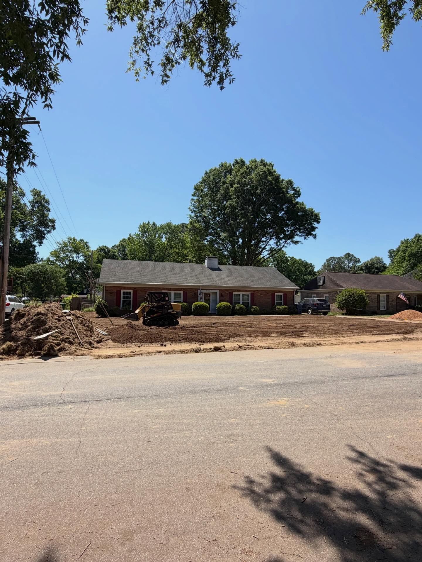 Single-story brick house across a road, with piles of dirt in front and trees under a clear blue sky