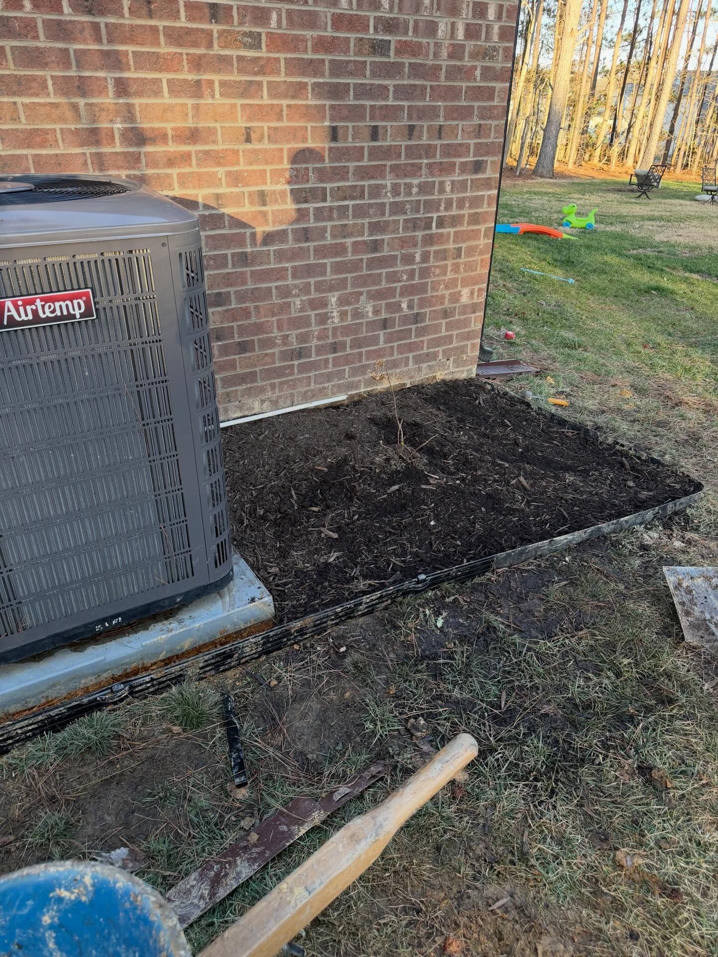 Mulch bed beside brick house with HVAC unit, shovel handle, and tools on grass