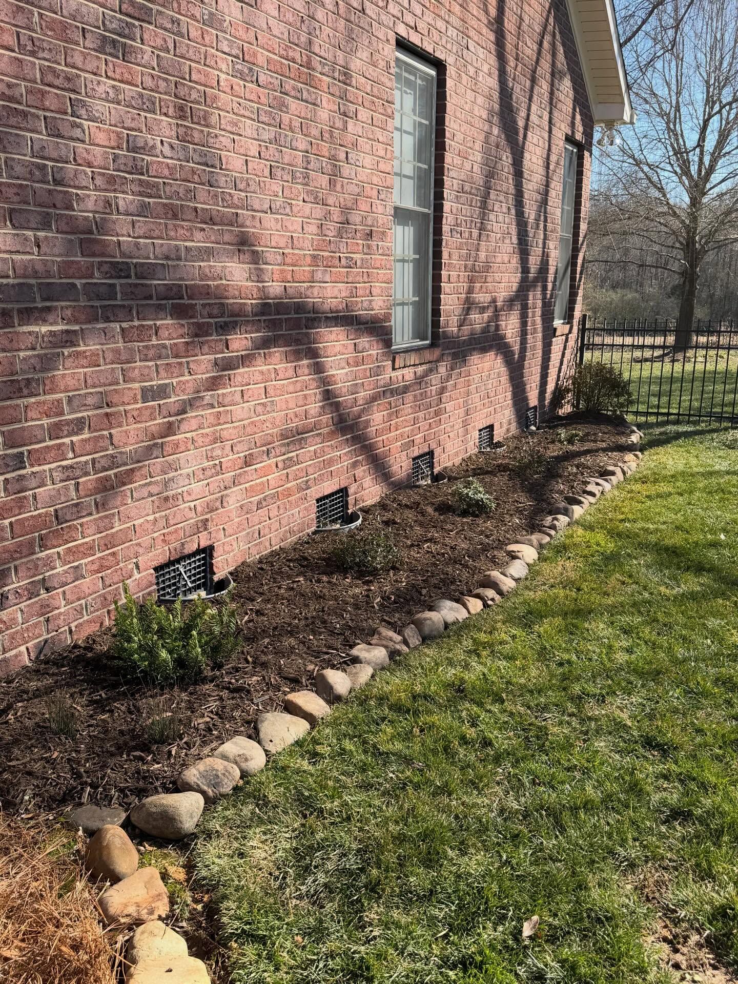 Brick house wall with a narrow garden bed of rocks and small plants along a grassy lawn.