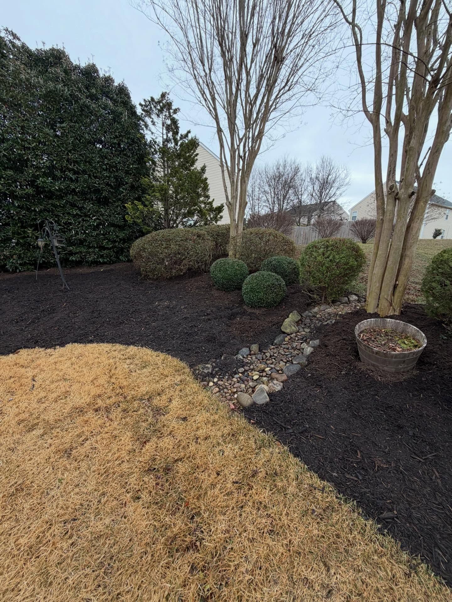 Landscaped yard with mulch beds, shrubs, bare trees, and a stone border beside a lawn.
