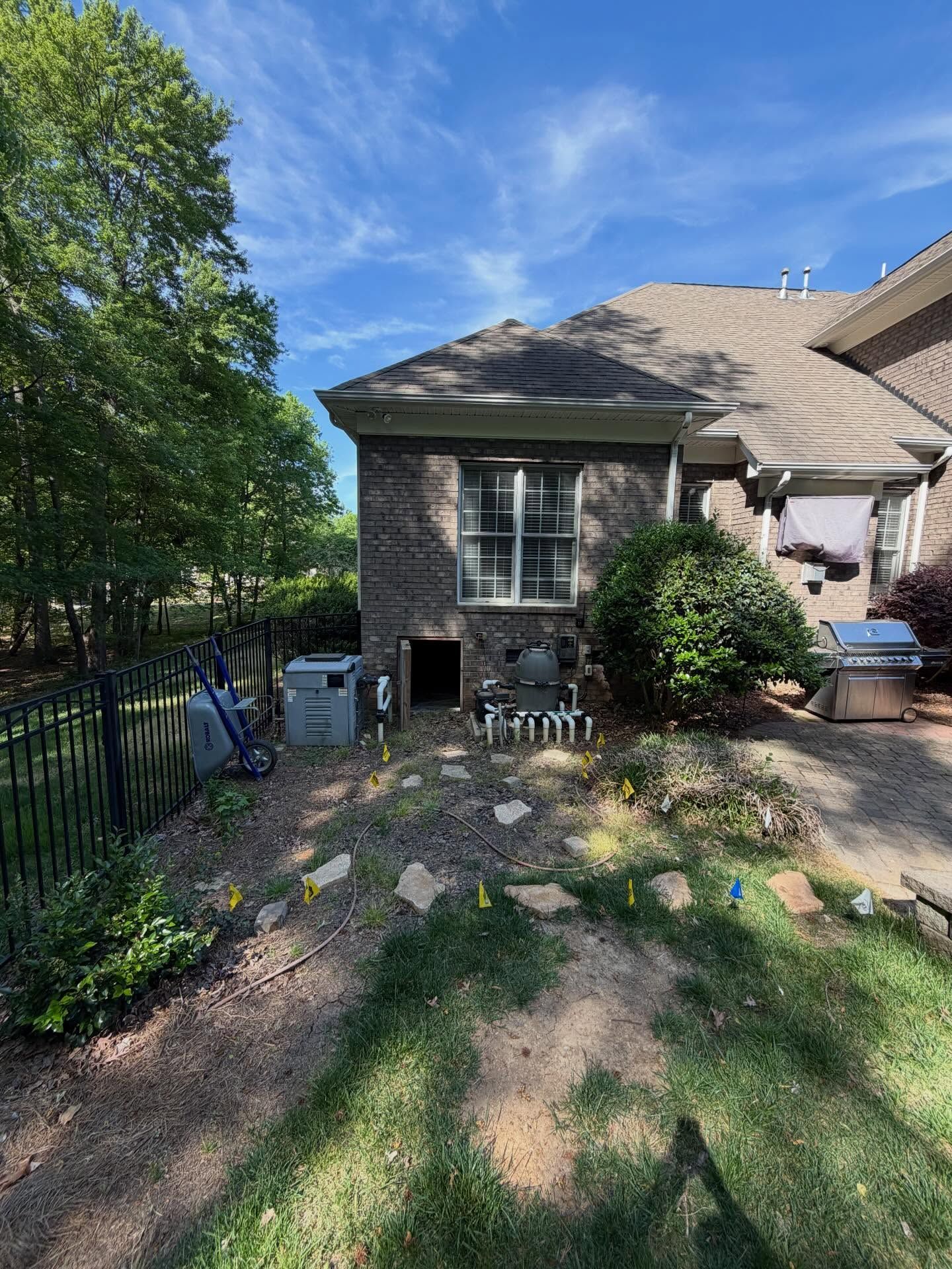 Backyard patio beside a brick house, with trees, shrubs, and a metal fence in bright sunlight