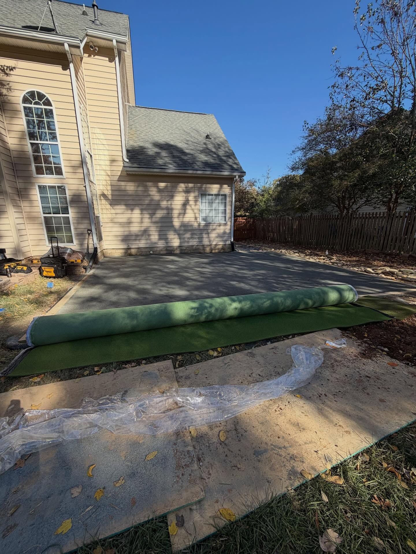 Backyard with a rolled green mat on a concrete patio beside a house and garage, surrounded by trees.