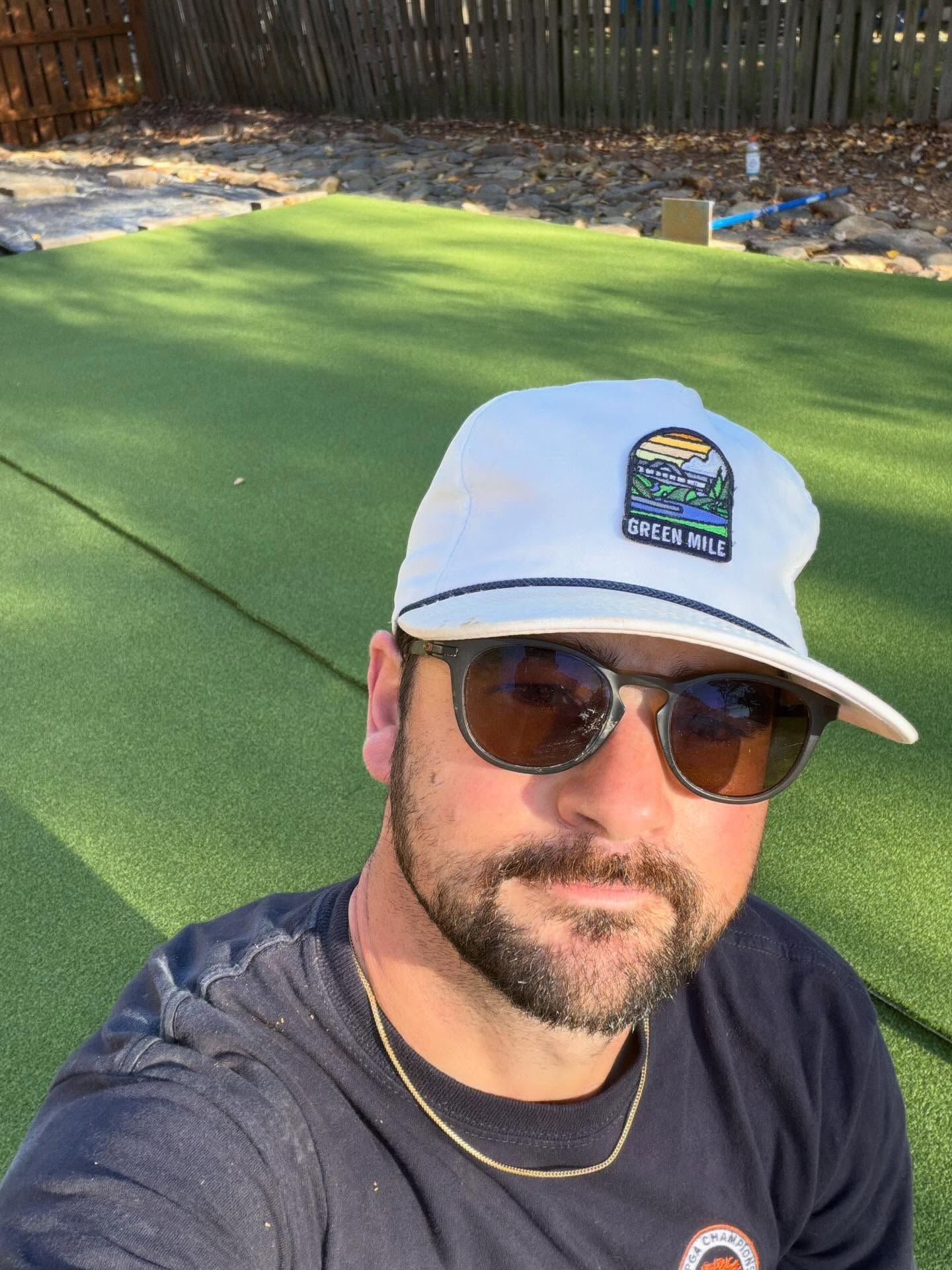Man wearing sunglasses and a white cap taking a selfie on a green backyard putting surface