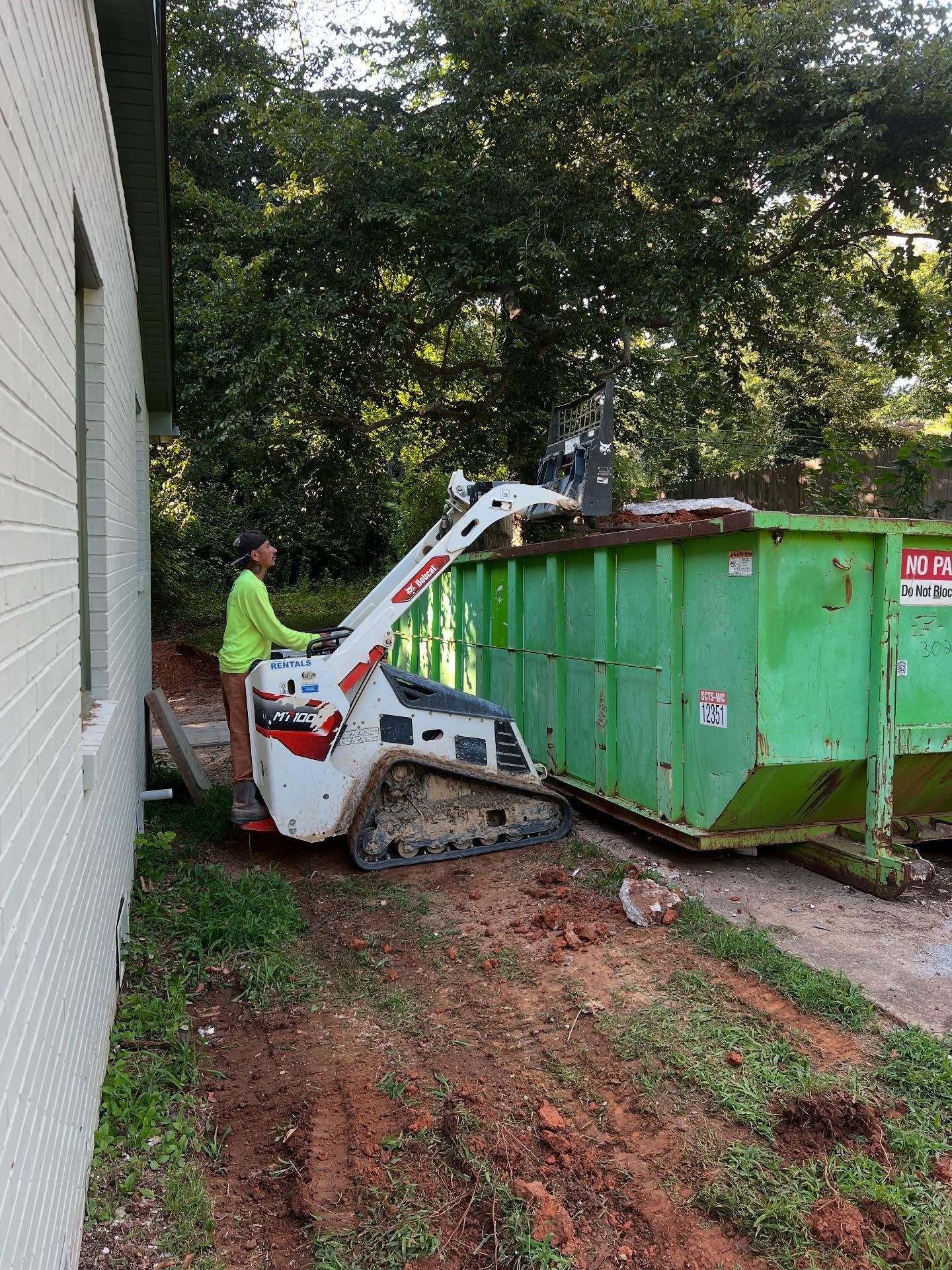 Worker operating a white skid-steer beside a green dumpster near a building and trees.
