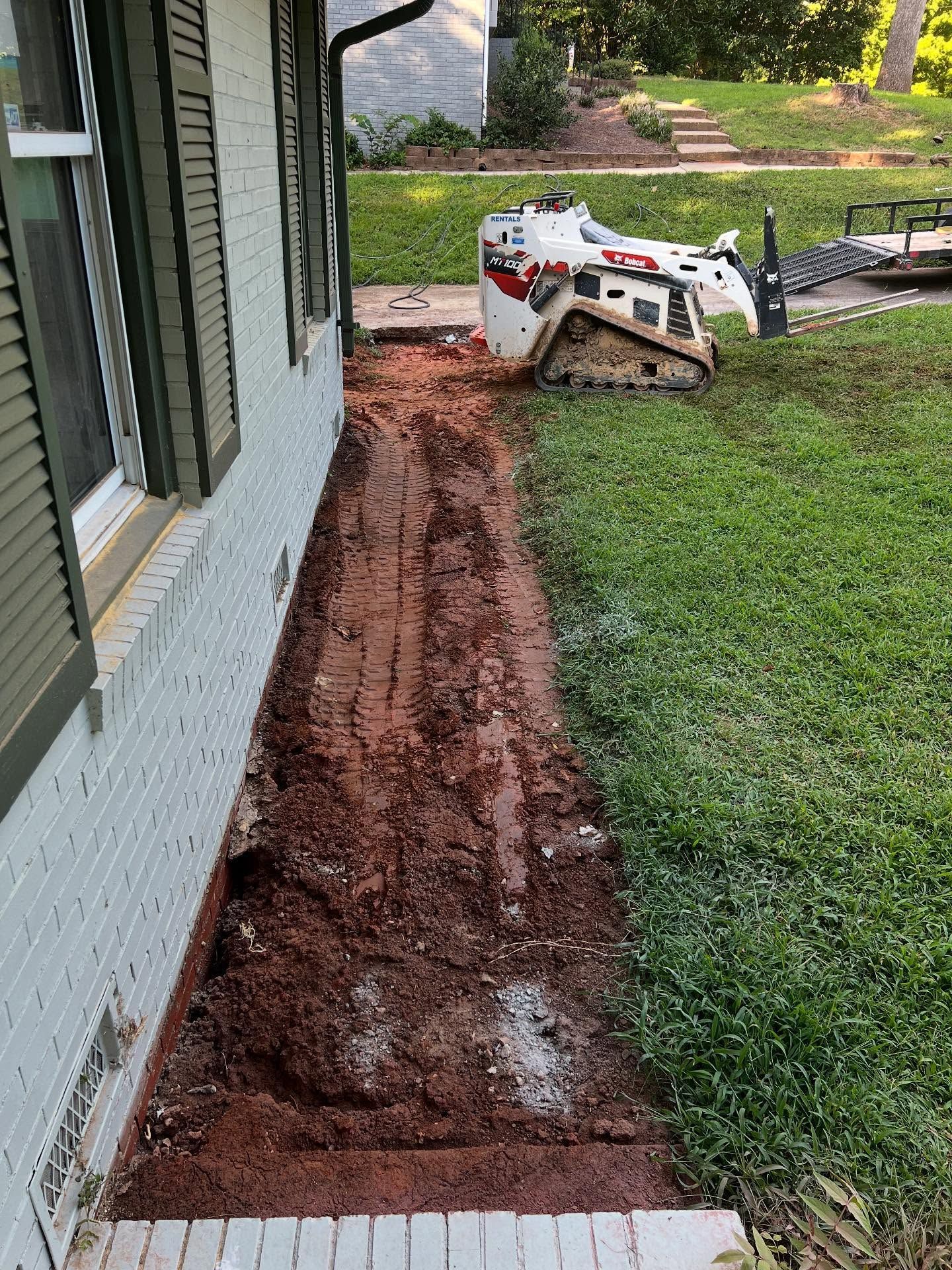 Freshly dug red-soil trench along a house foundation, with a small skid steer on the lawn.