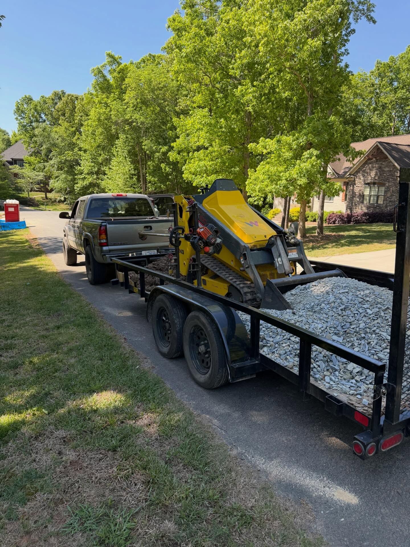 Truck hauling a yellow skid steer on a dual-axle trailer in a leafy neighborhood driveway