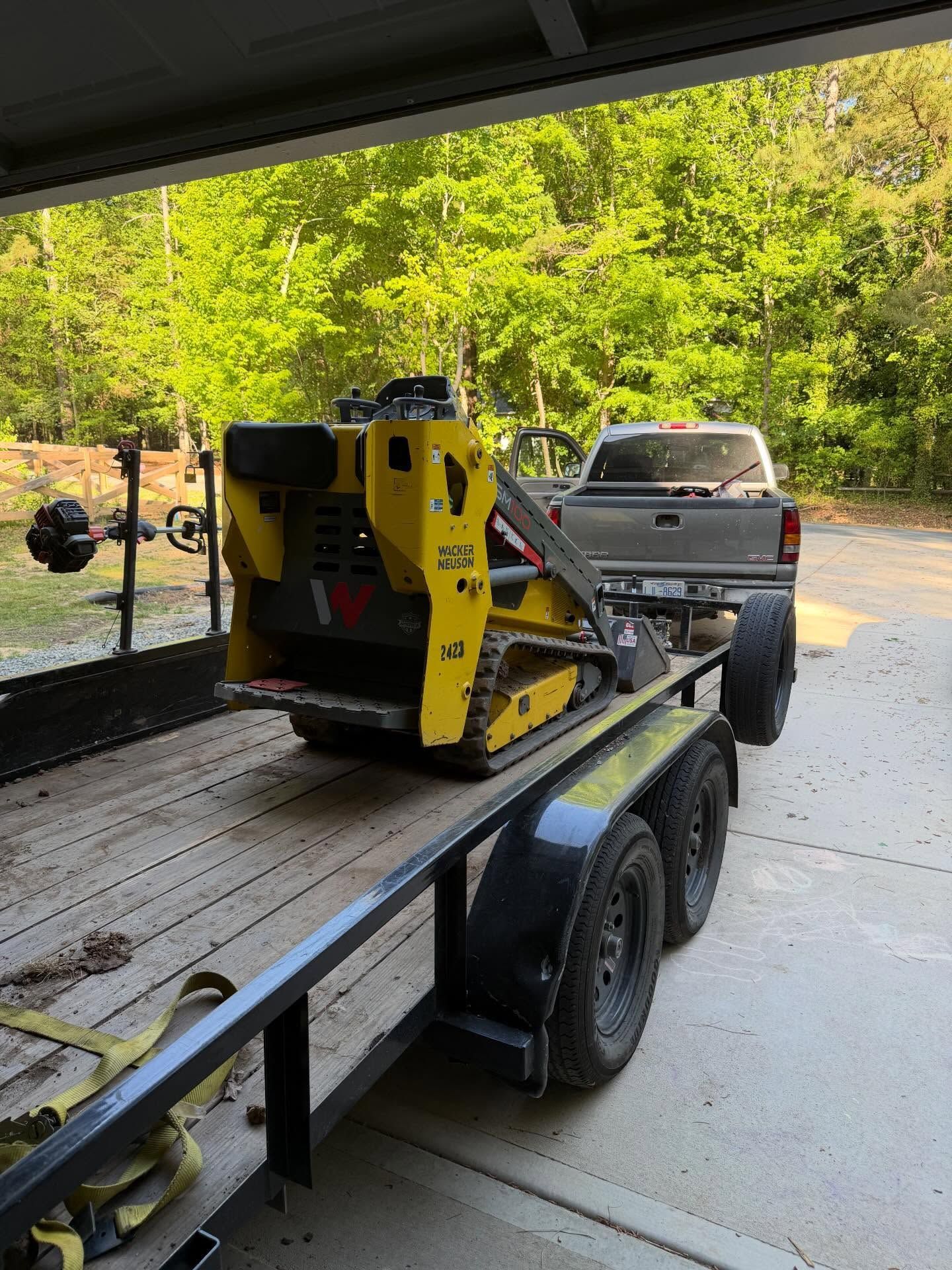 Yellow construction machine on a flatbed trailer being unloaded in a garage, with a pickup truck nearby.