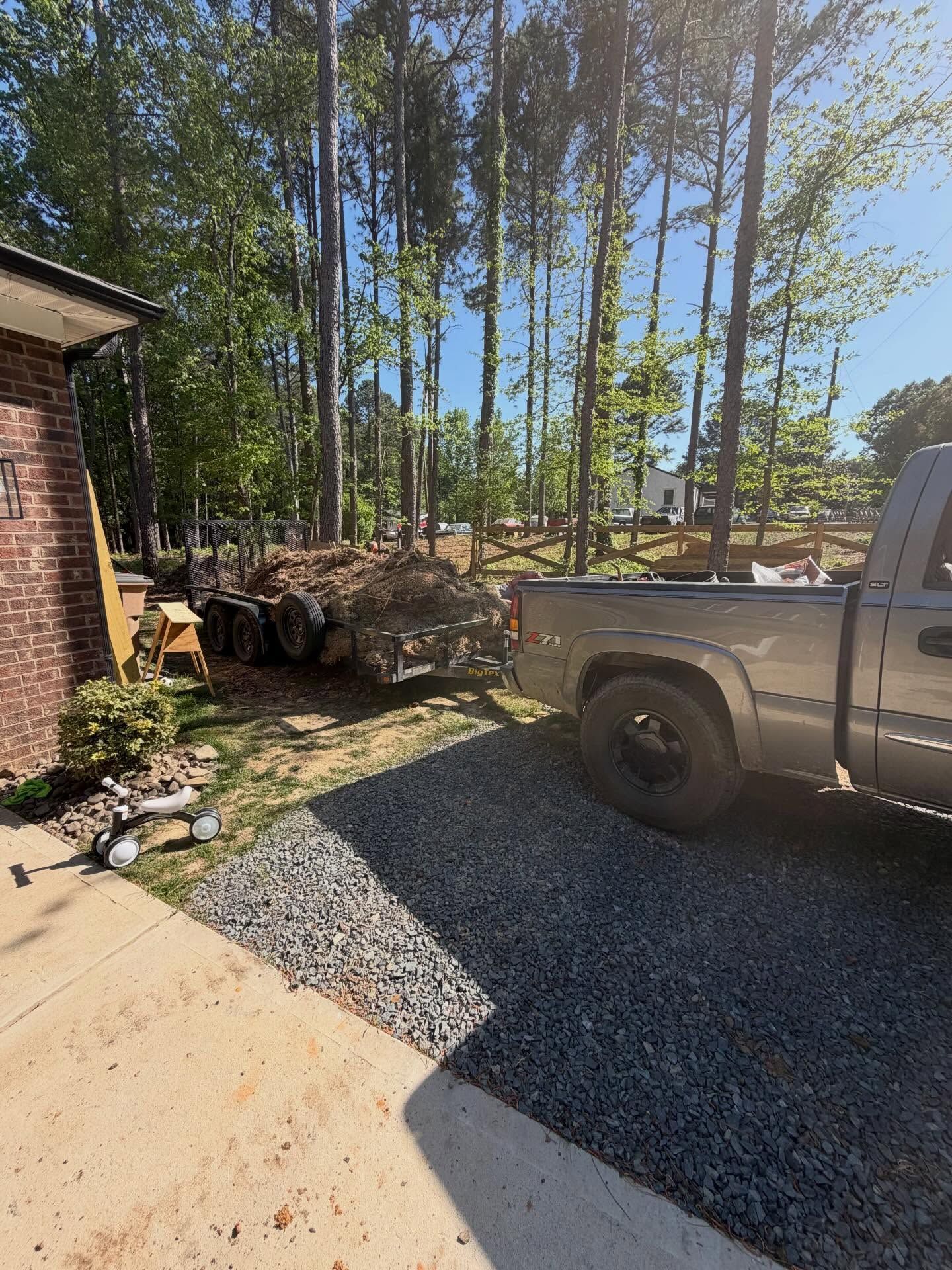 Pickup truck parked on gravel beside a brick house and stacked firewood under tall trees