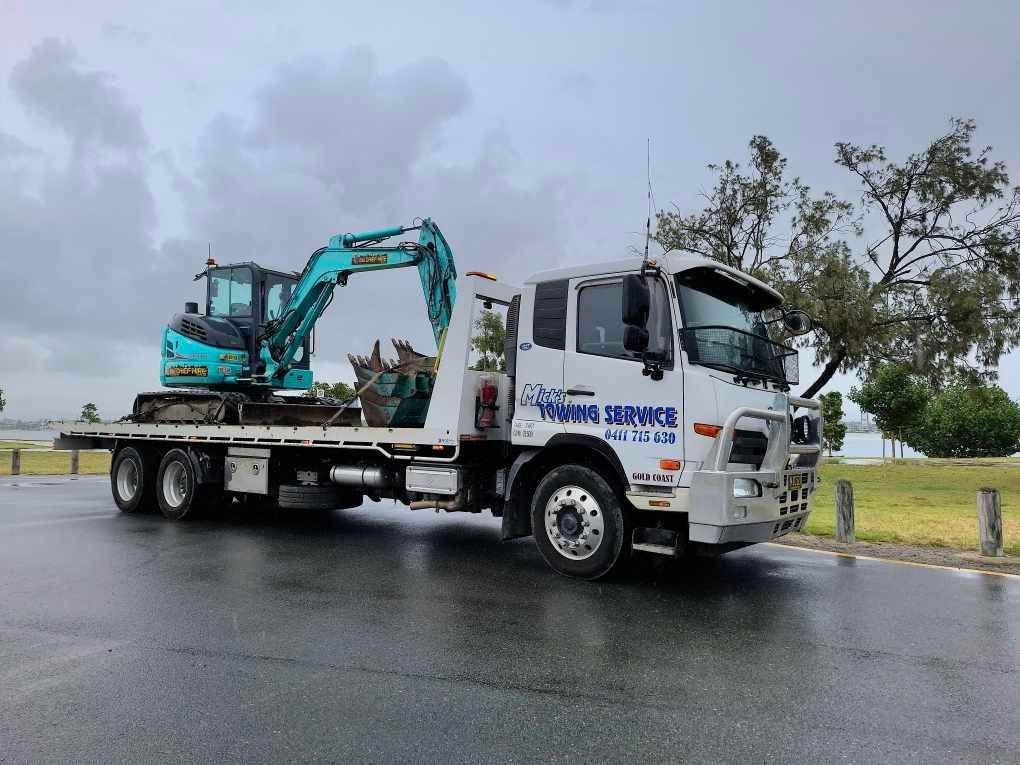 A Tow Truck With an Excavator on the Back is Parked on the Side of the Road — Mick's Towing Service Pty Ltd in Carrara, QLD