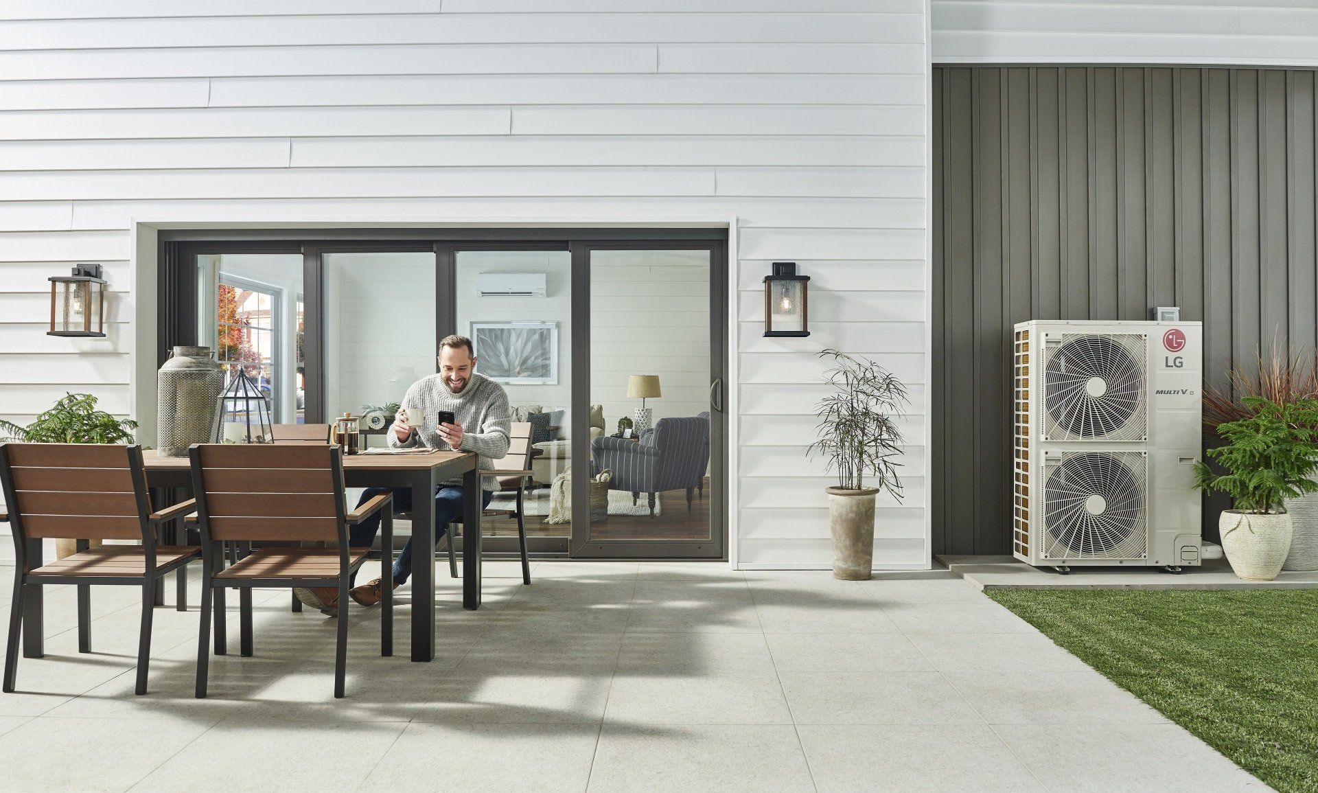 Man at outdoor table, near sliding glass door, looking at a phone, with a heat pump visible.