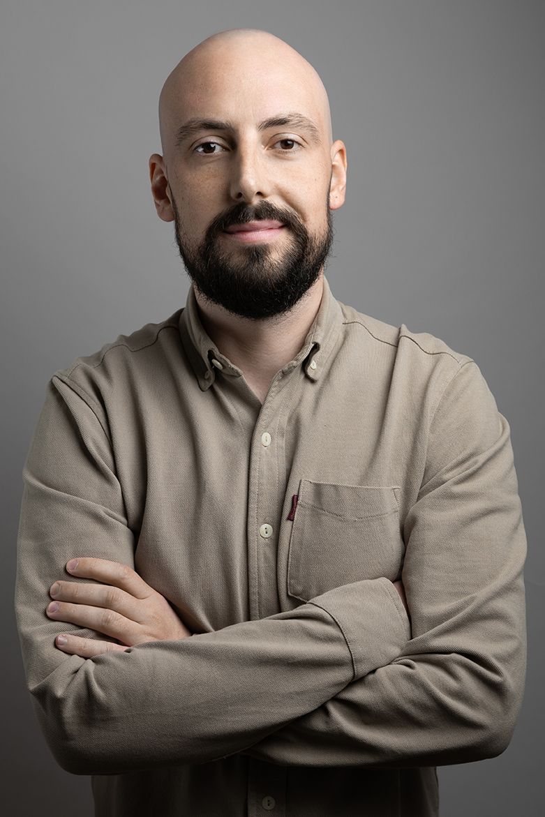 Un homme chauve avec une barbe est debout, les bras croisés.