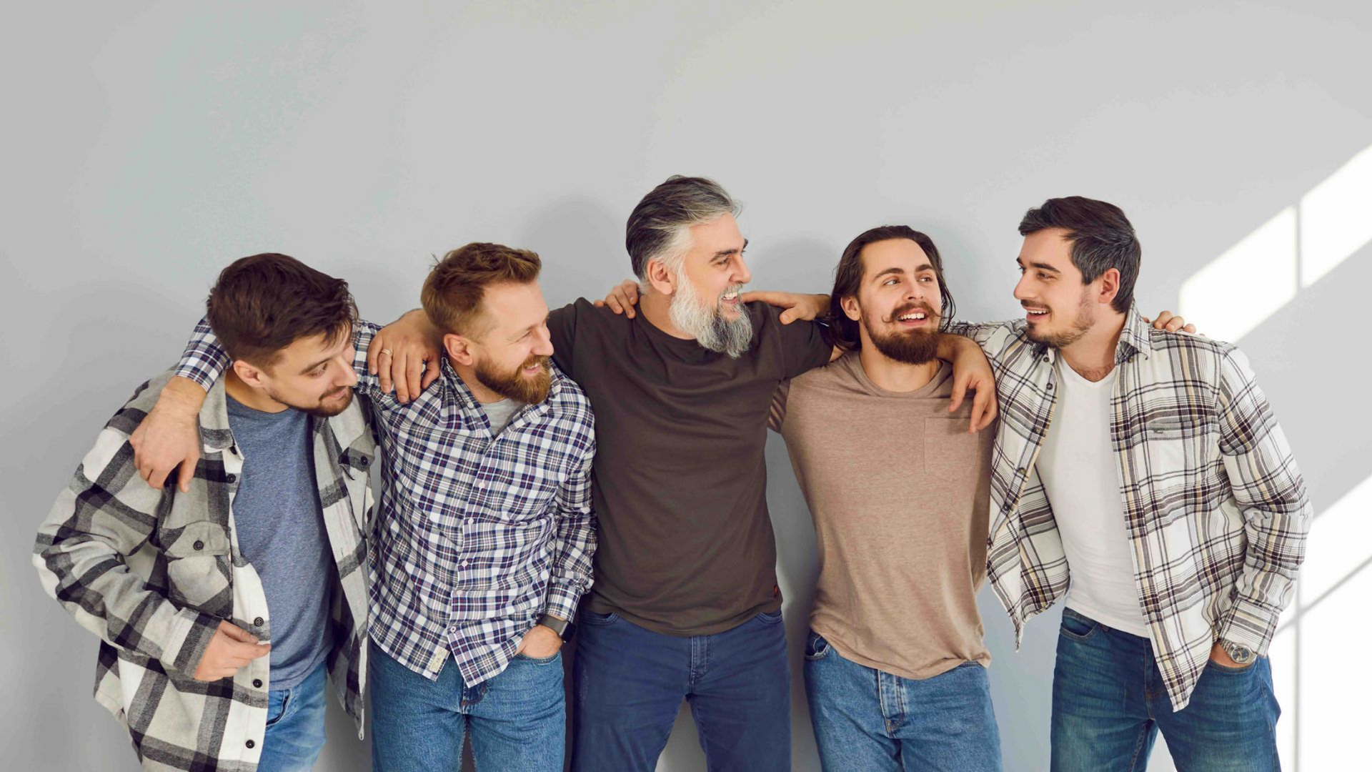 Four friends stand arm-in-arm, smiling in casual clothes against a light gray wall.